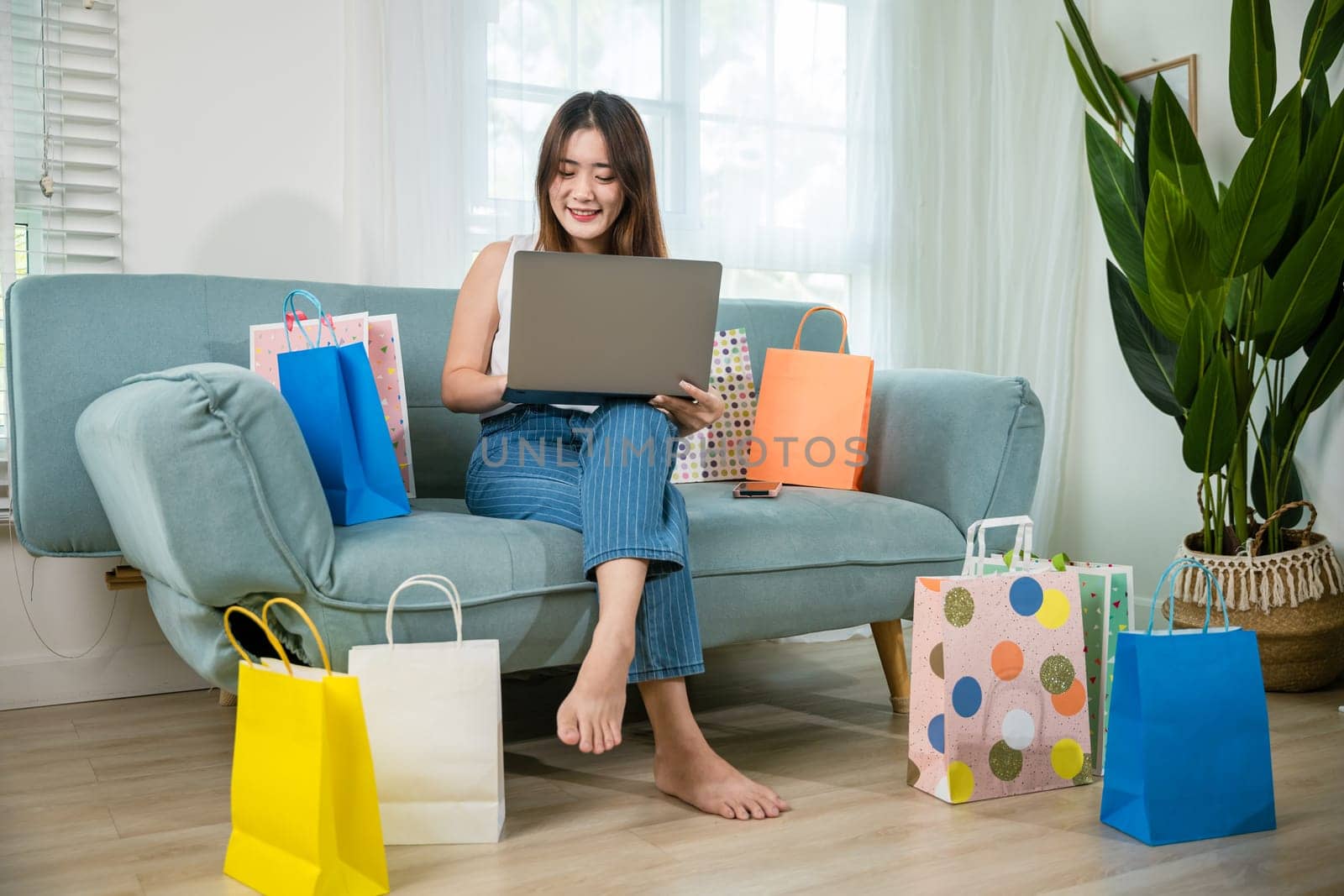 Happy woman customer buyer sitting on sofa with shopping bags using laptop computer to shop online in living room at home, Asian young female satisfied customer with purchase on web site, shopping day