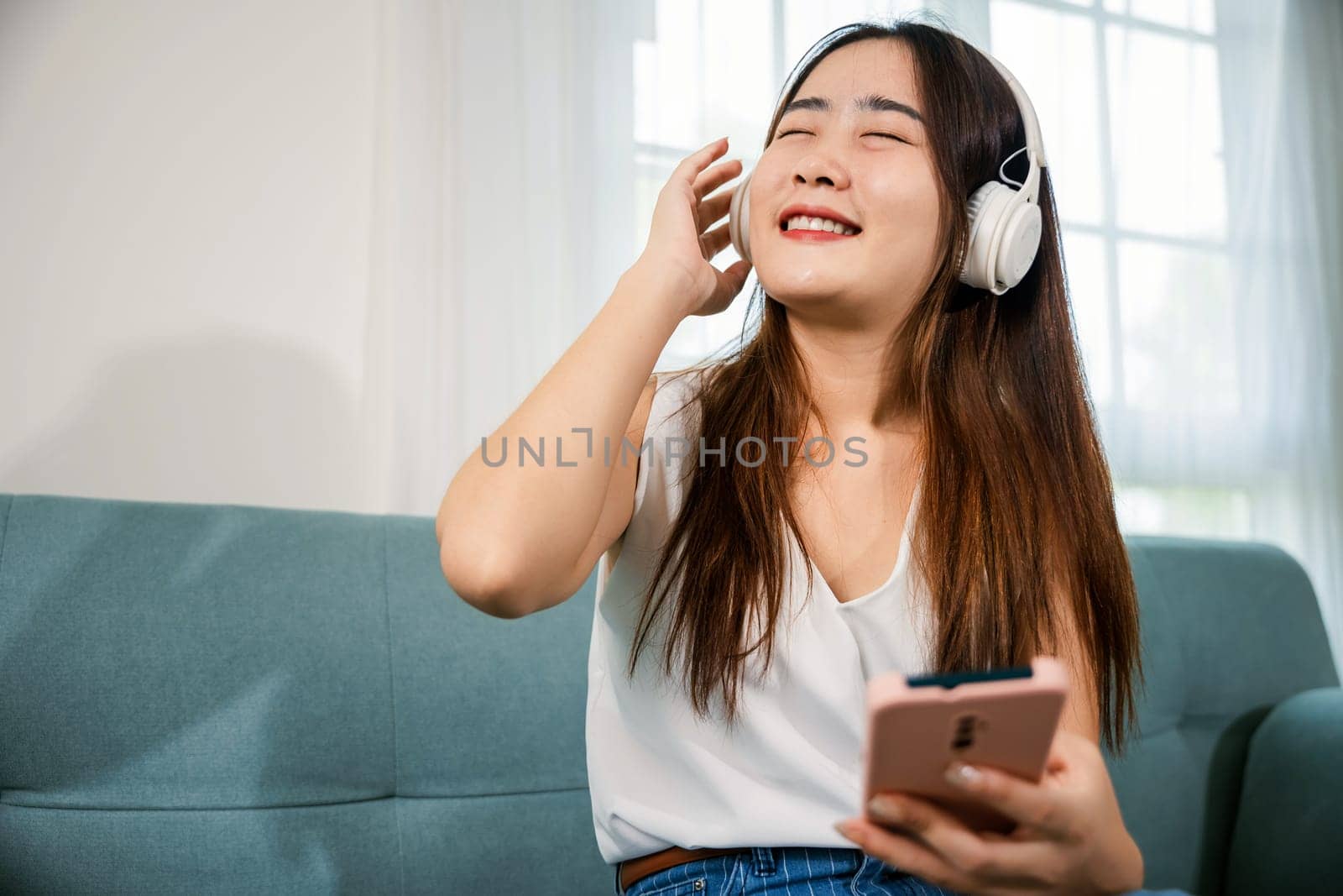 Asian young woman wearing headphones listening to music with smart mobile phone and relaxing while sitting on sofa in living room at home, female wear earphone listen music on smartphone
