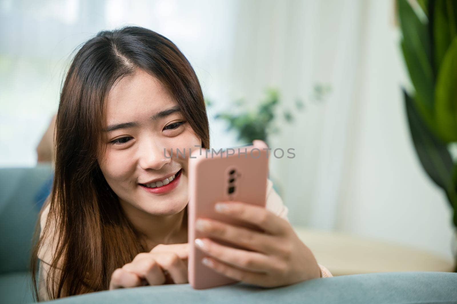Smiling attractive woman relaxed reading text message on smartphone, Asian young female using smart mobile phone lying on sofa in living room room at home, gadget online application making internet