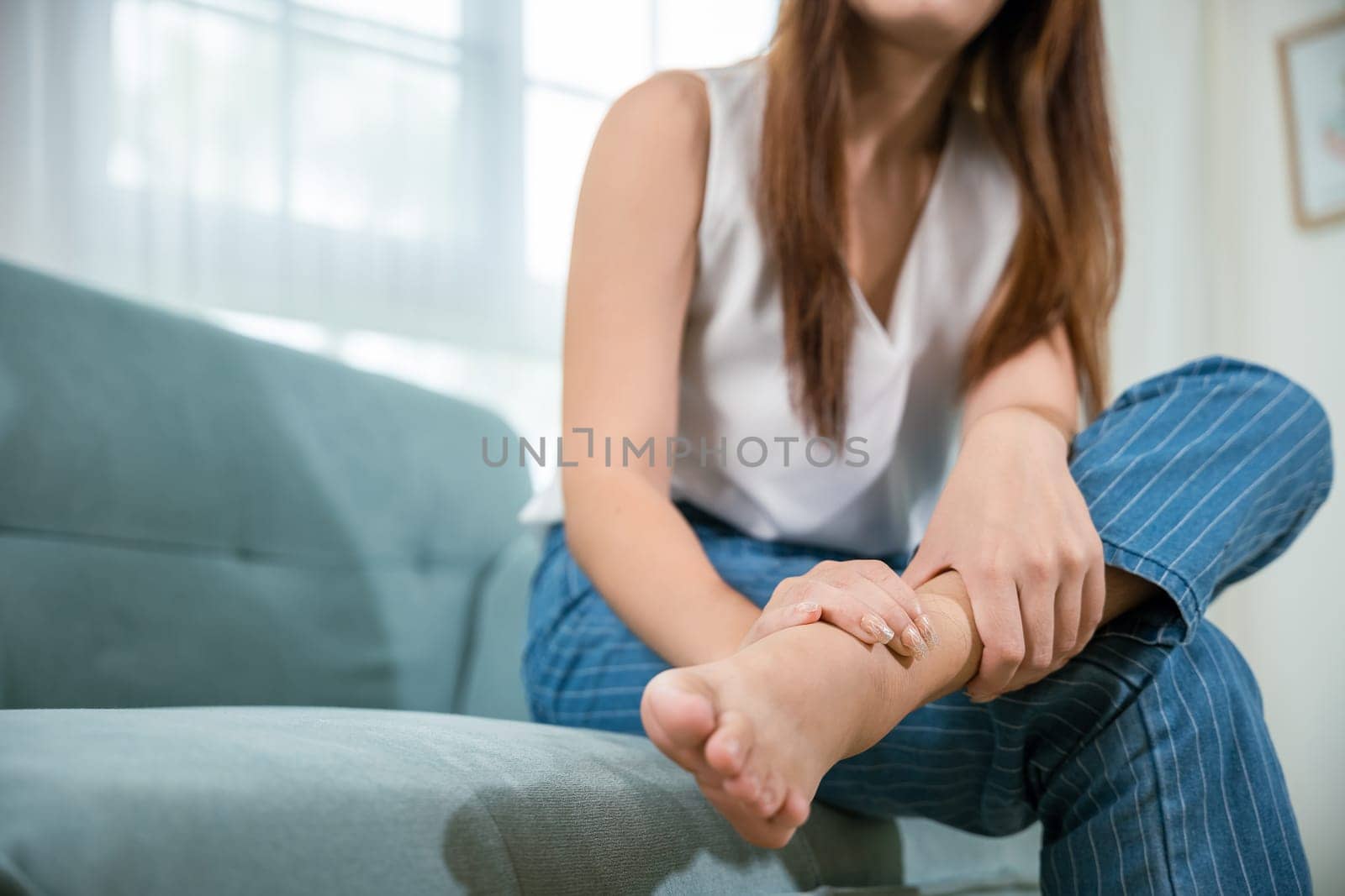 Asian beautiful female problems with foot at home, painful ankle injury, Young woman sitting on sofa holding her feet and stretch muscles have symptoms feeling pain, Health care and medical concept