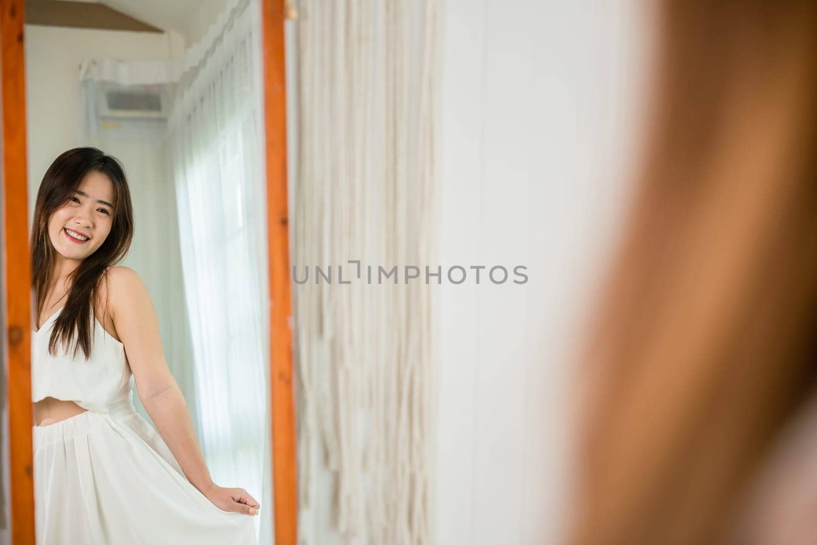 Happy woman wearing dress on front mirror looking clothes at home after morning wake up, Asian beautiful young female trying on blouse and smiling while standing in front of mirror in room