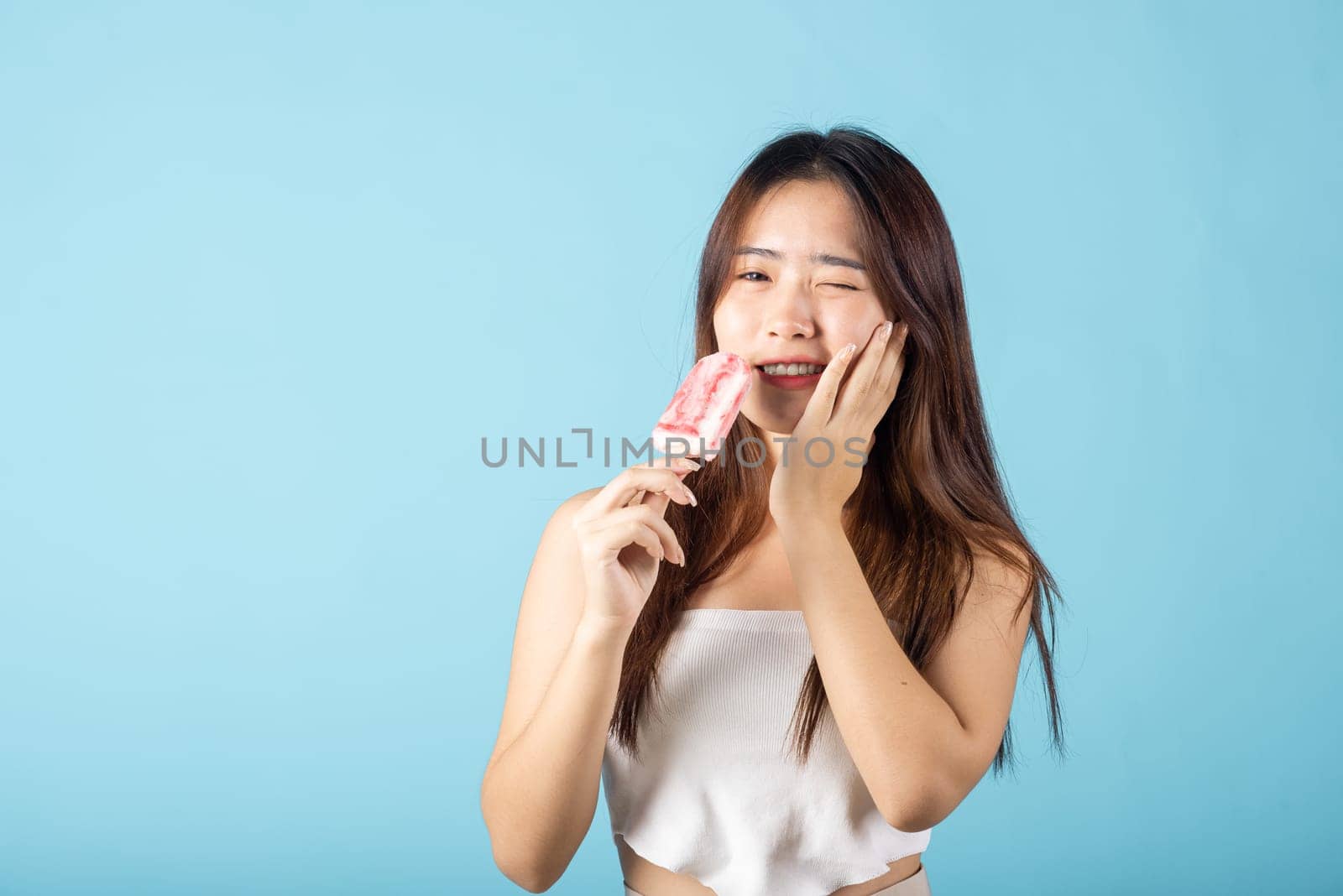 Portrait of Asian young woman with sensitive teeth after eating delicious ice cream wood stick mixed fruit flavor feeling painful uncomfortable, studio shot isolated on blue background, dental problem