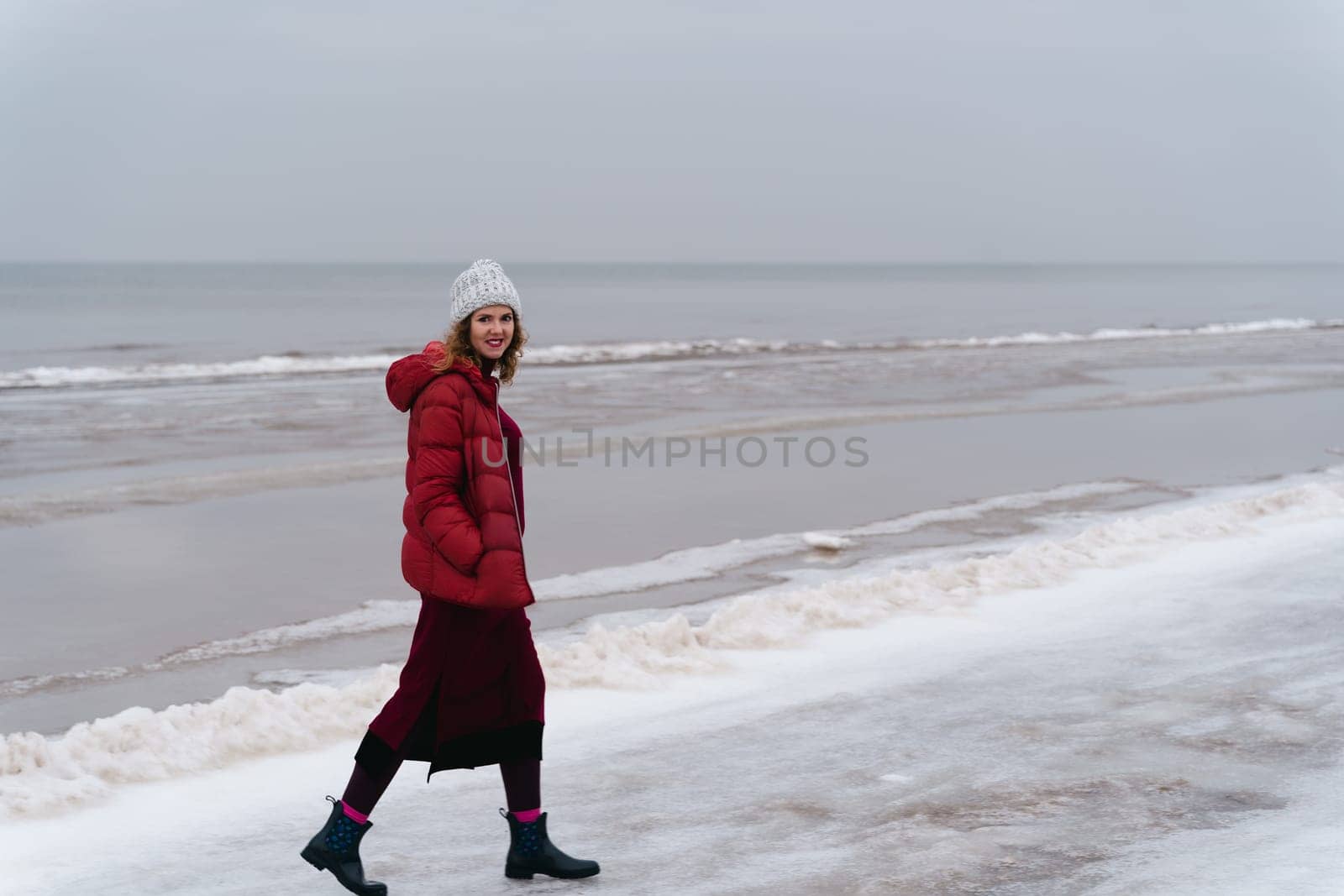 A woman in red warm clothes walks along the sea in winter.