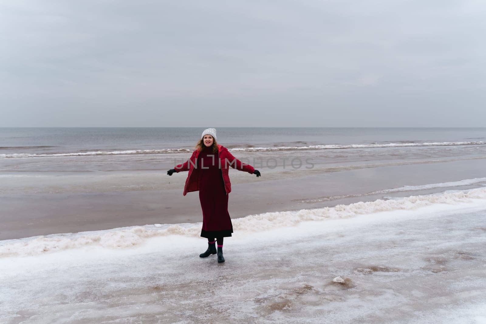 A joyful woman in a red jacket and a dress by the sea in winter.