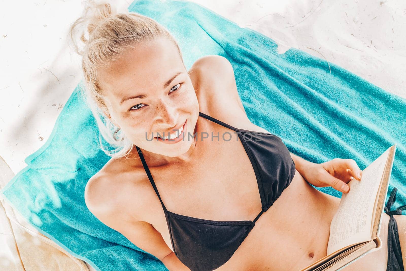Young beautiful blonde caucasian woman in black bikini enjoying reading book on the beach while sunbathing.