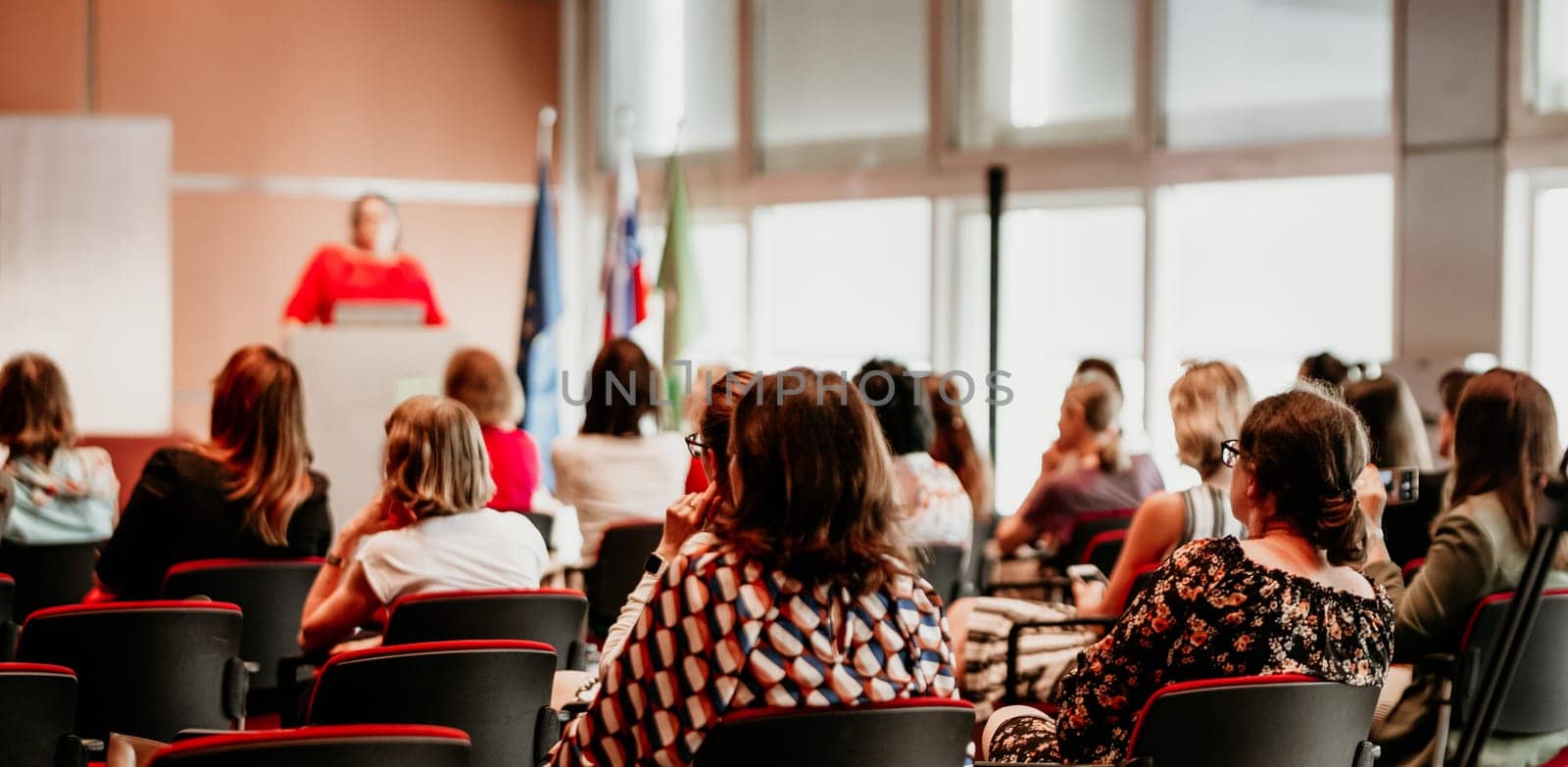 Business and entrepreneurship symposium. Female speaker giving a talk at business meeting. Audience in conference hall. Rear view of unrecognized participant in audience.