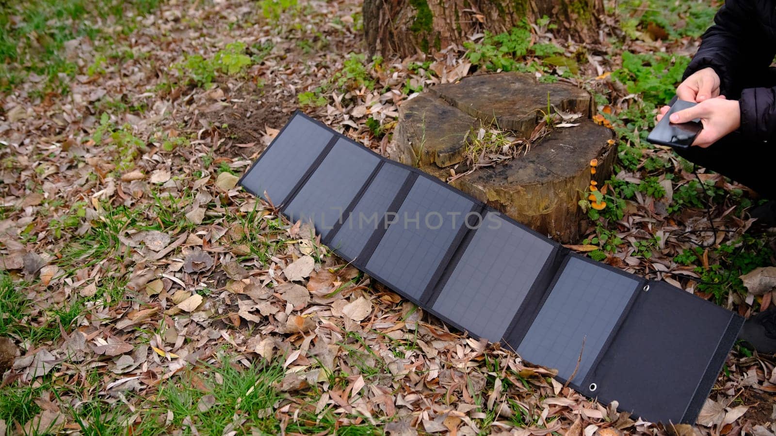 Hands plug in portable solar panel charging smartphone by mutlipurpose cable, power bank, watches, laptops nature background. Clean energy for using in camping or at home when NO electricity