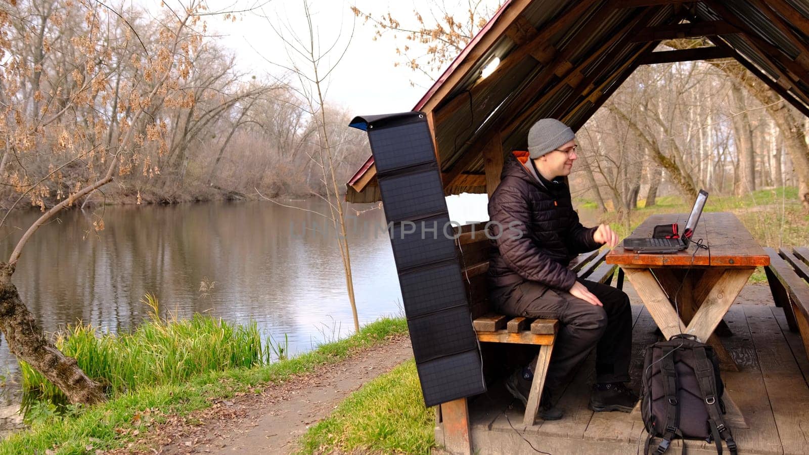 Man work on laptop charginf from portable solar panel utdoor in alclove near the river. Clean energy for using in camping. Work with gadgets outdoor with portable solar panels