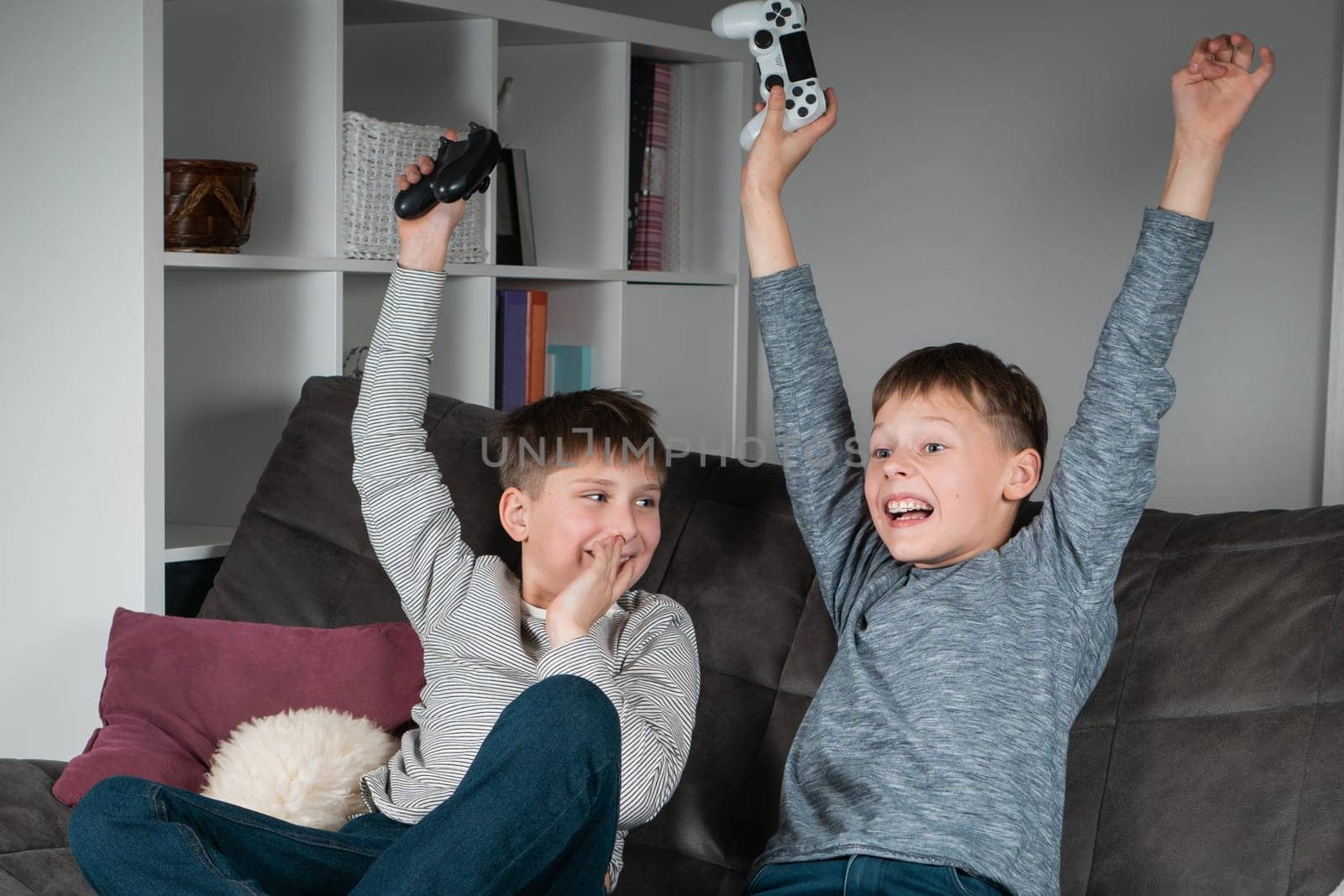 Portrait of two wonderful positive teenage boys sitting on grey sofa at home, holding gaming controller joystick, playing videogames, celebrating, raising hands, winning competition. Hobby, free time.