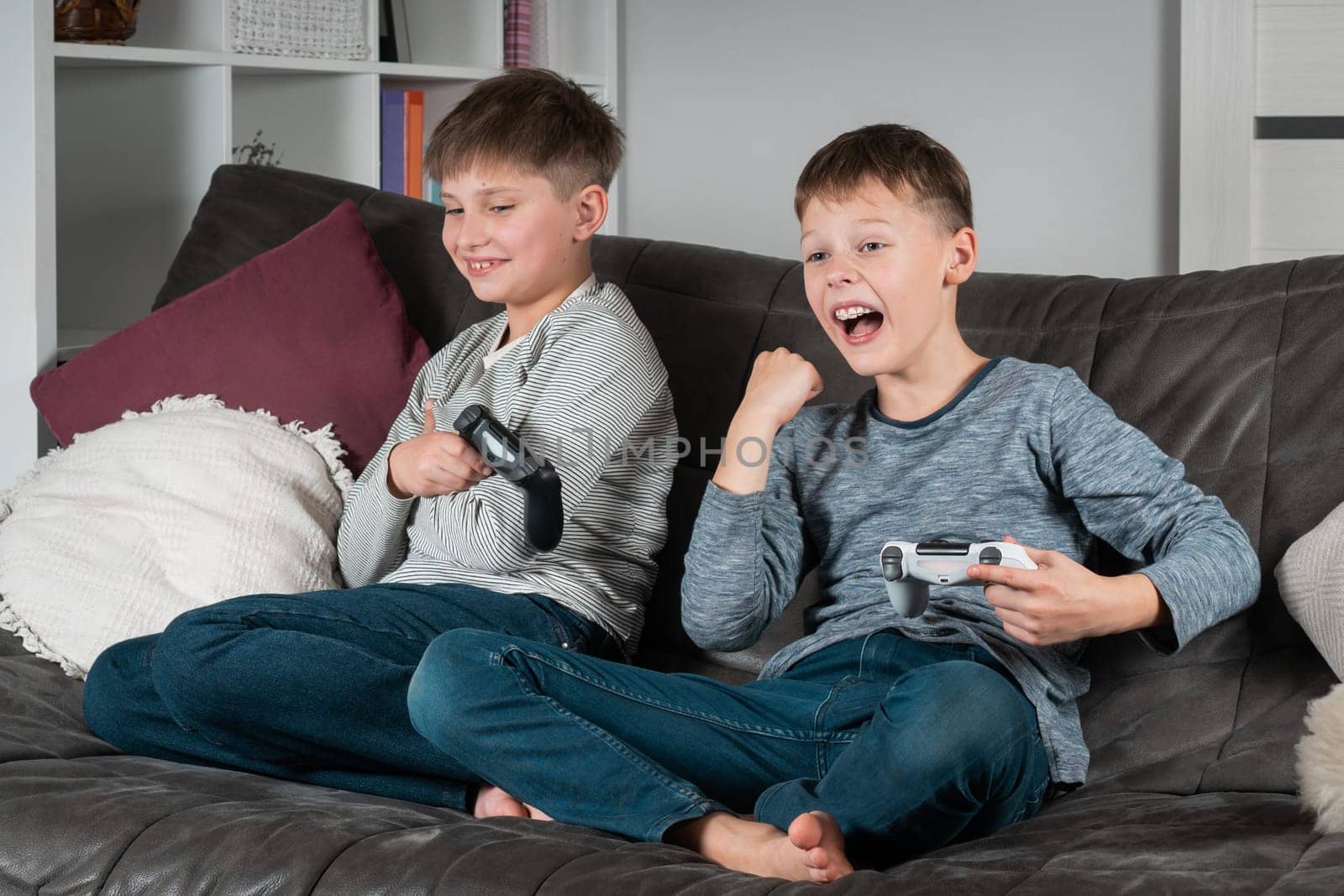 Portrait of two happy teenage boys sitting on grey sofa at home with open mouth, holding gaming controller joystick, playing videogames, screaming, raising hand, celebrating victory. Hobby, free time.