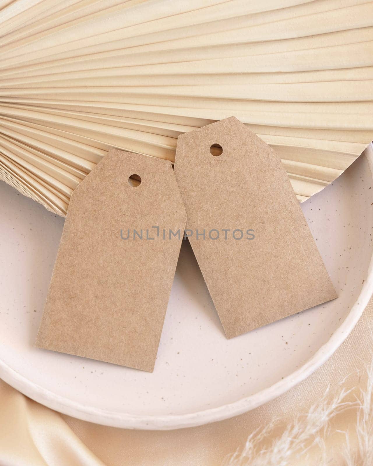 Blank gift tags on beige satin fabric and dried palm leaf close up, mockup. Boho scene with kraft paper envelope. Bohemian or close to nature greeting or wedding Invitation. Romantic composition.