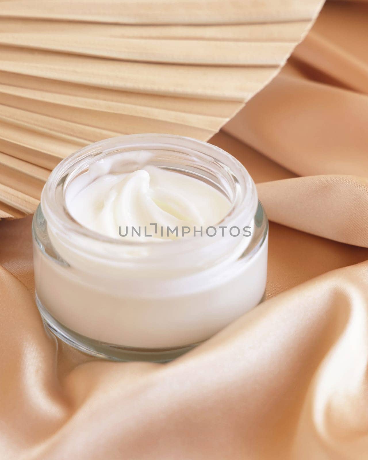 Opened cosmetic jar on beige satin fabric and dried palm leaf close up, mockup. Boho scene with glass cream jar. Bohemian or close to nature scene.