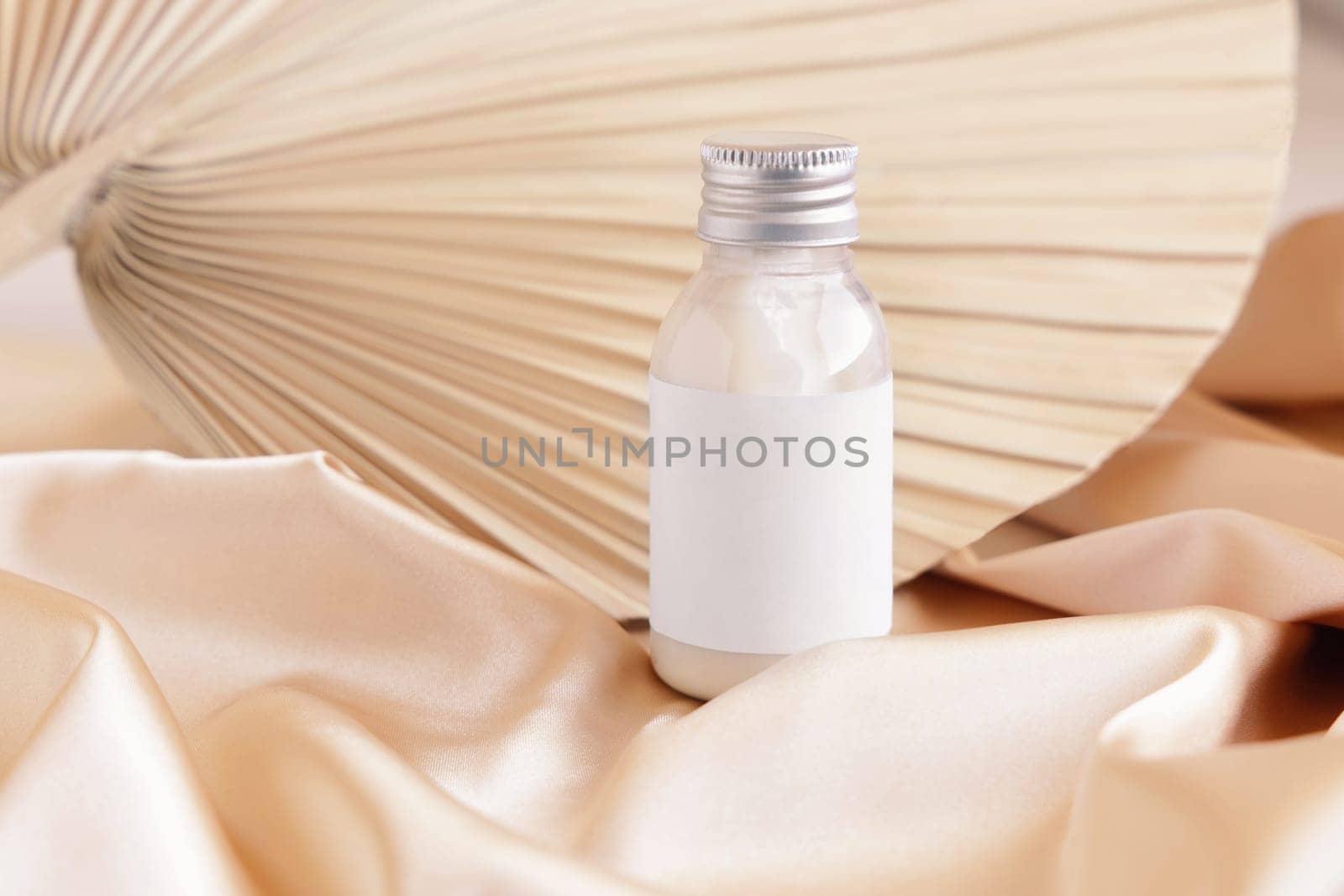 Cosmetic bottle with blank label on beige satin fabric and dried palm leaf close up, mockup. Boho scene small cream bottle. Bohemian or close to nature composition.