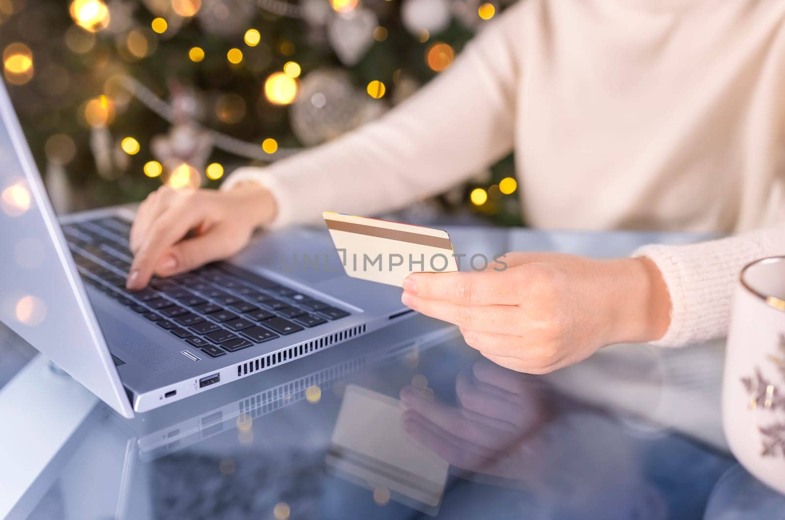 Cropped shot of female hands with golden credit card and laptop. Christmas presents, decorated tree on background. Woman shopping for holiday season presents online. Wooden table, copy space, close up