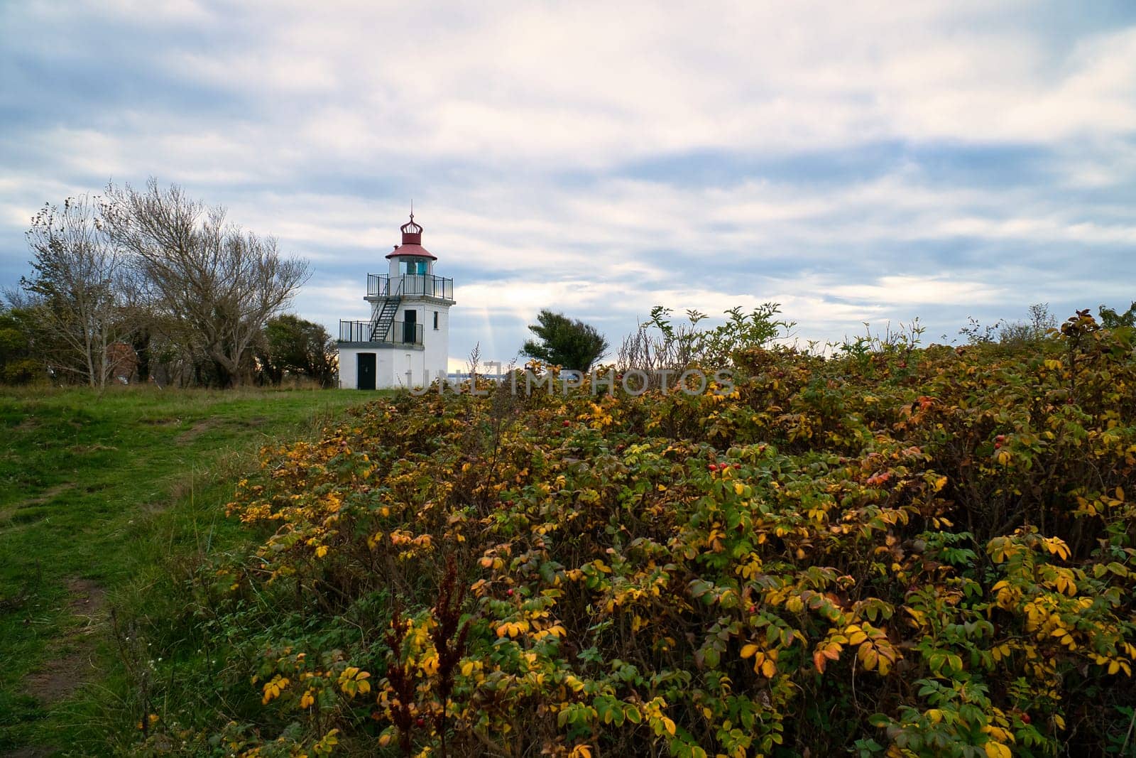 Lighthouse, Spodsbjerg Fyr in Huntsted on the coast of Denmark. Sun rays shining through the clouds. Meadow with trees. Landscape photo from sea
