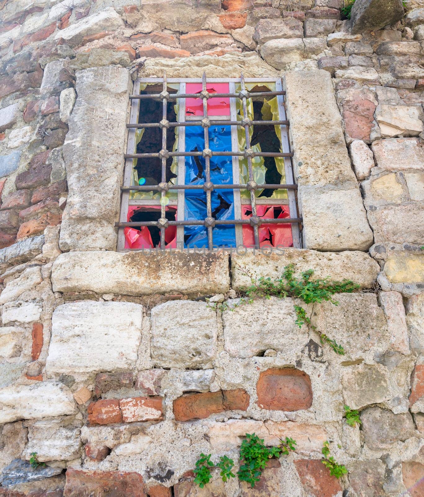 Abandoned weathered old stone bricks wall with corroded antique grunge wrought iron small window with multi vibrant colored squares, in an old village