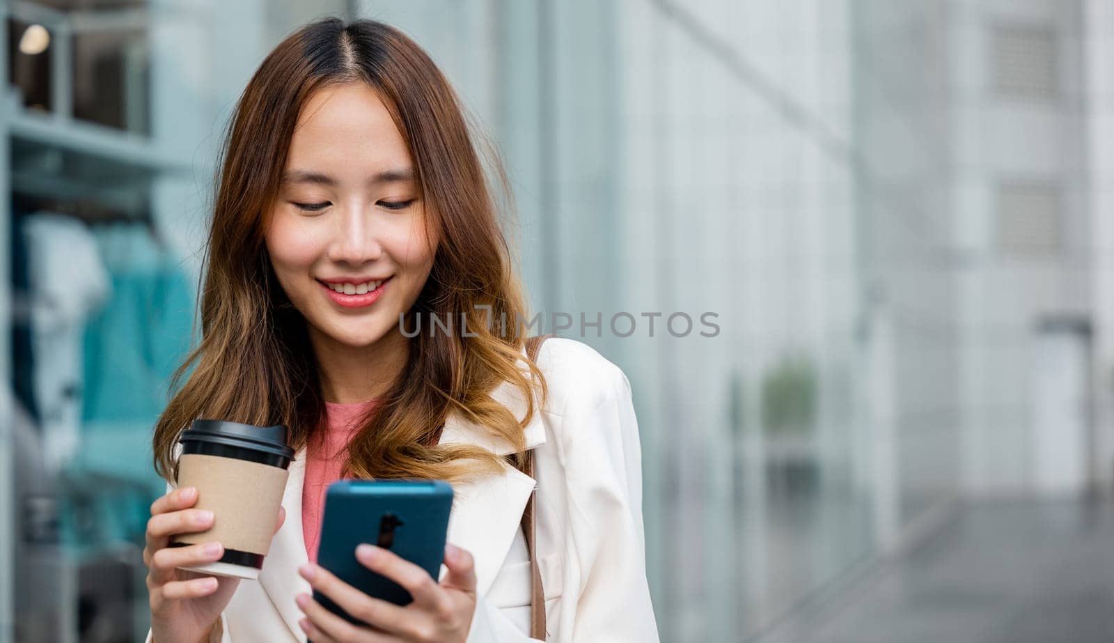 Happy business woman hold mobile phone and paper cup of hot drink outdoor walking on city street, Asian businesswoman holding coffee cup takeaway and smartphone go to work she walking near her office