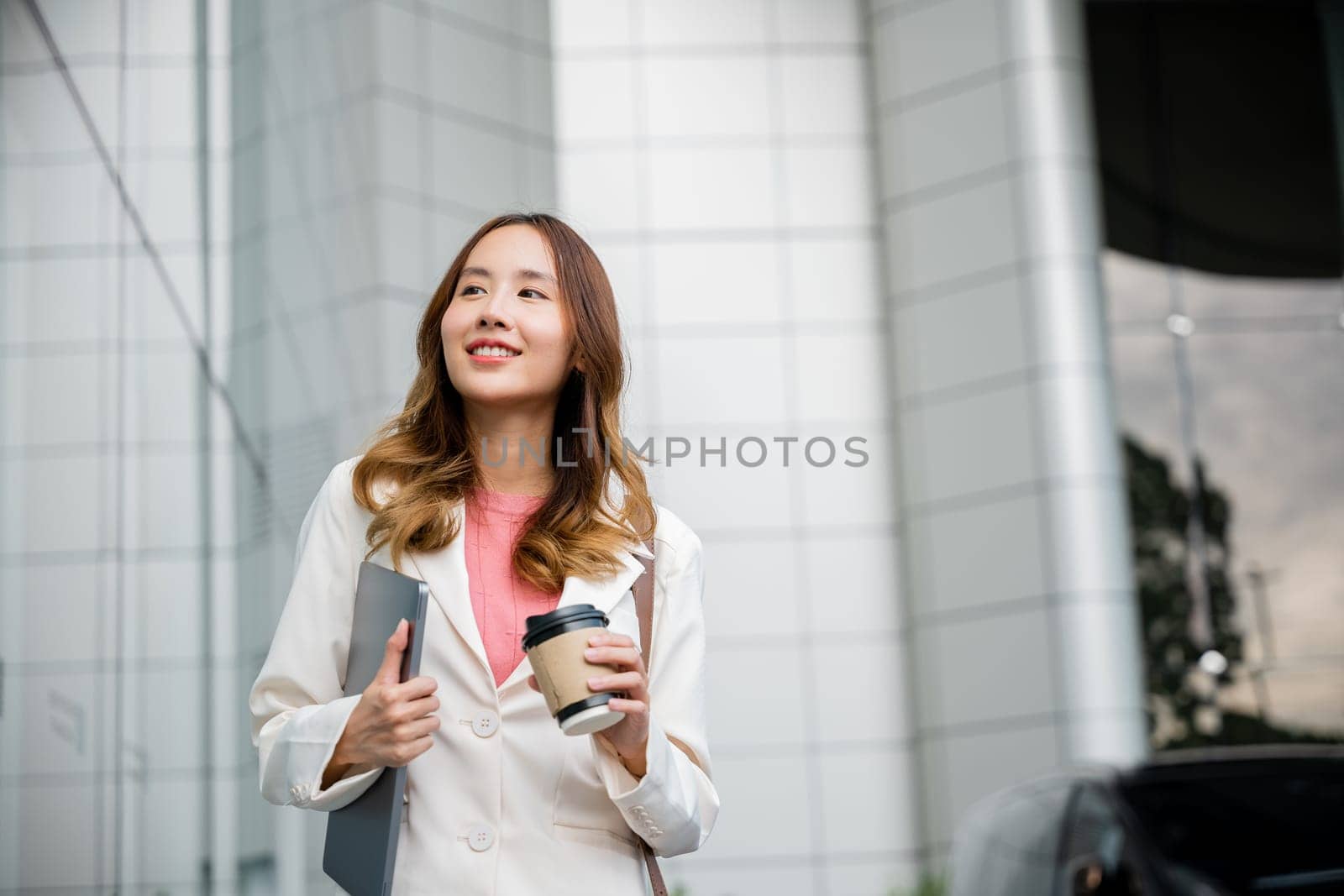 Young business woman wearing white suit jacket and smile while go to work outdoor mirror building background. Confident Businesswoman with cup of coffee holding Laptop walking outside office building