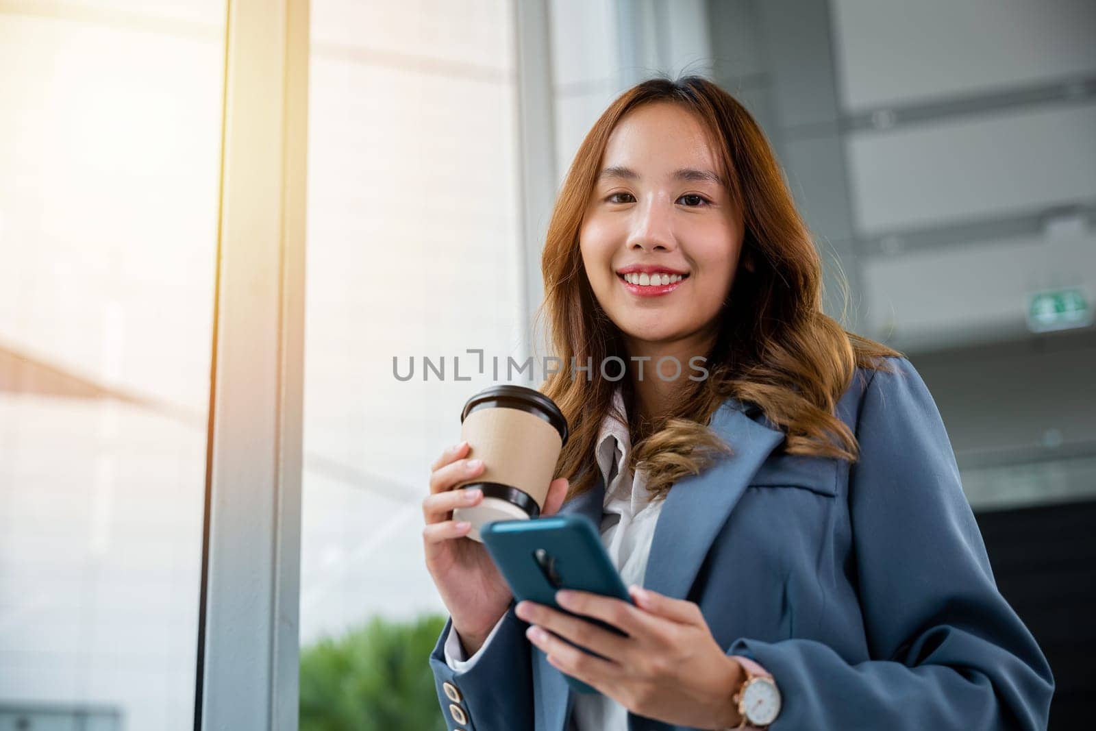 Young Asian businesswoman wearing blue shirt holding coffee cup and her smartphone. business woman smiling standing holding mobile phone near window and elevator blur background in the office.