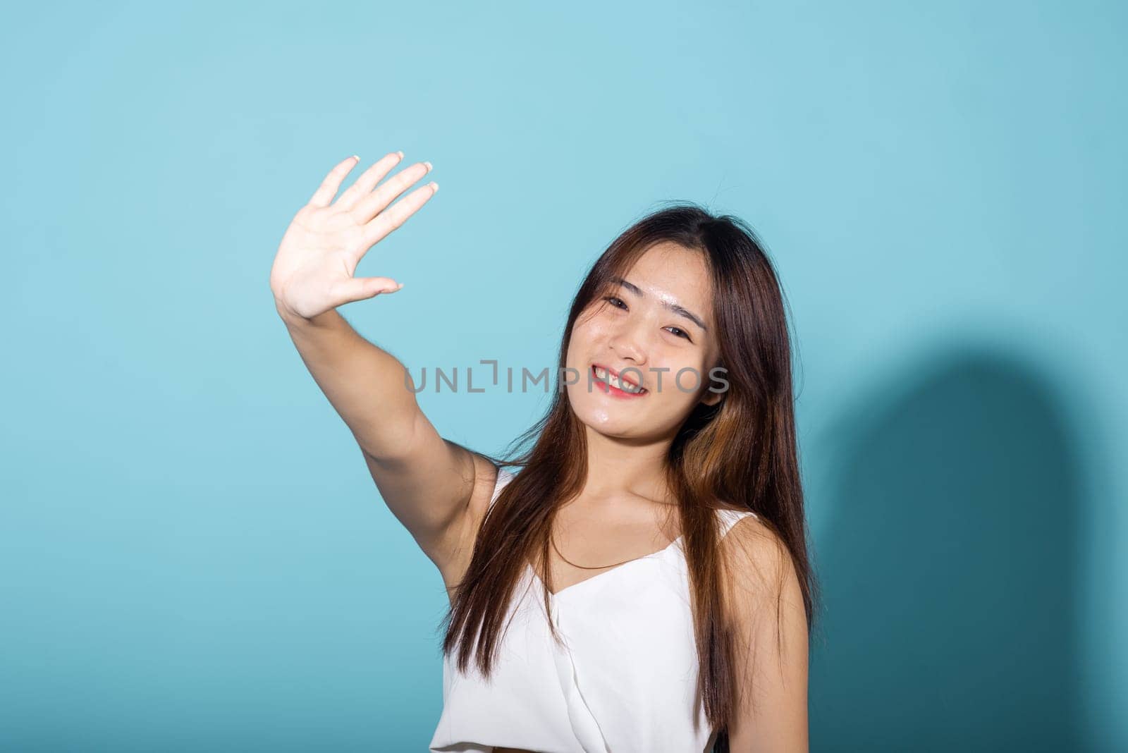 Female raising arm and hand in protection and block sunshine in studio shot isolated on blue background, Asian young woman smile hand up cover face protect sun light and UV, sunscreen cream advertise