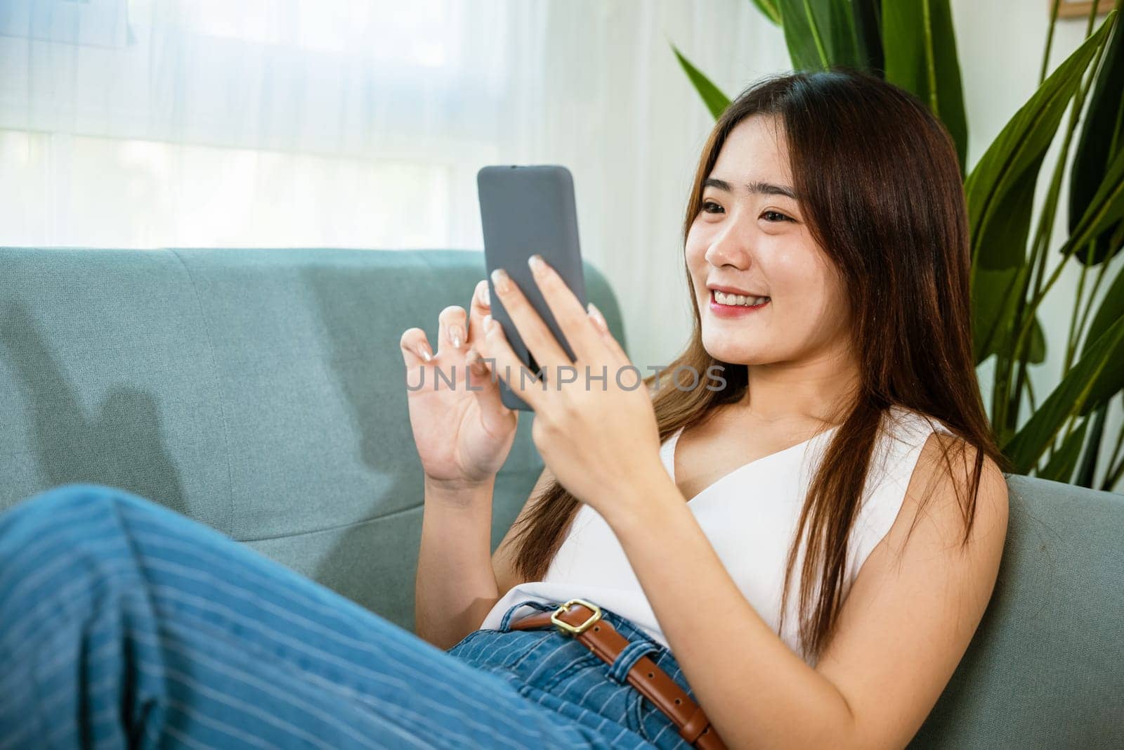 Happy Asian young woman checking social media holding smartphone at home while relaxing on sofa living room, Smile beautiful female sitting on couch using mobile phone to shopping online