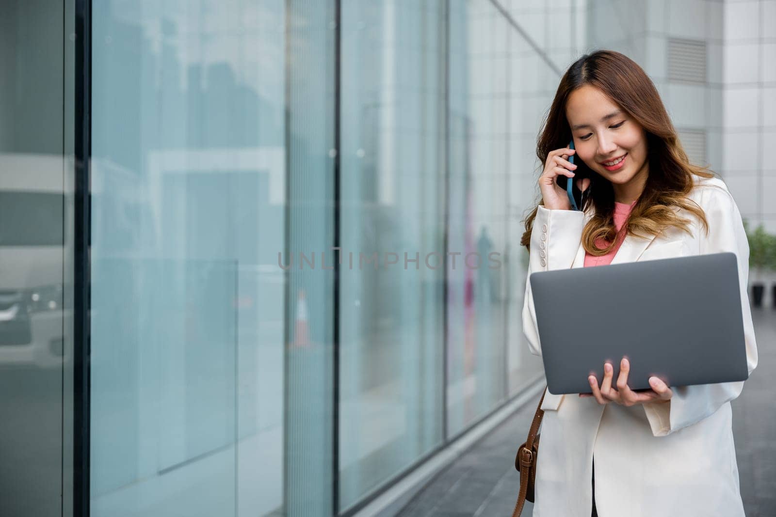 Asian businesswoman working on laptop and talking on cell phone at front building near office, Portrait beautiful business woman in white suit smile discussing issues on smart mobile phone in city