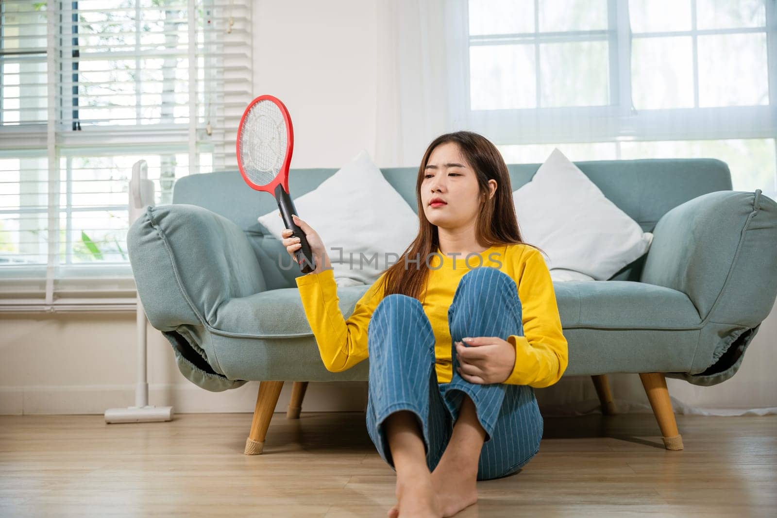 Woman killing mosquitoes hand holding fly swatter like weapon in living room at home, Asian young female sitting floor using mosquito swatter or electric net racket, technology insect killer