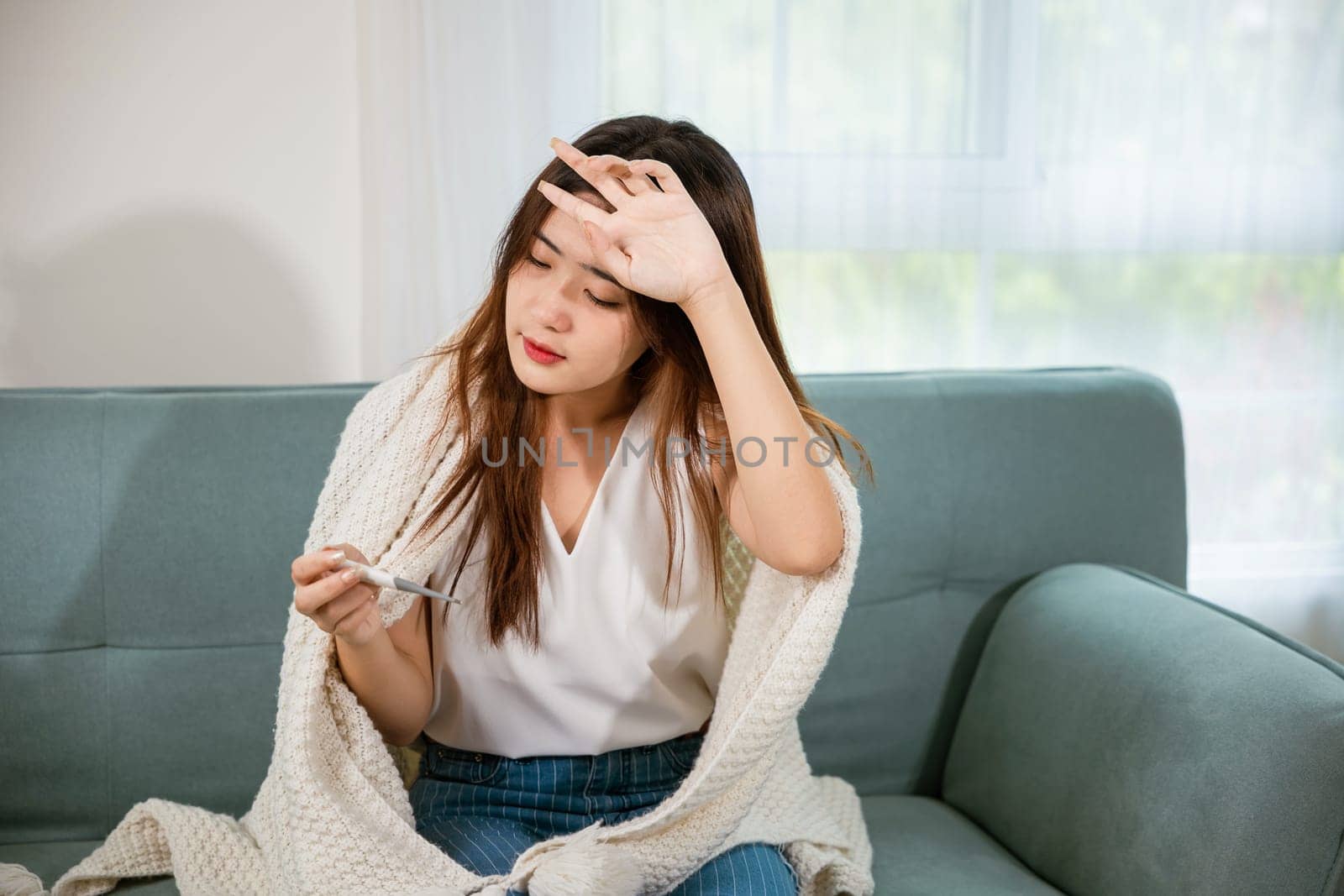 Sick Asian young woman sit under blanket on sofa she sneeze checking temp with digital thermometer and tissue paper in living room at home, Cold flu and migraine concept, medical healthcare