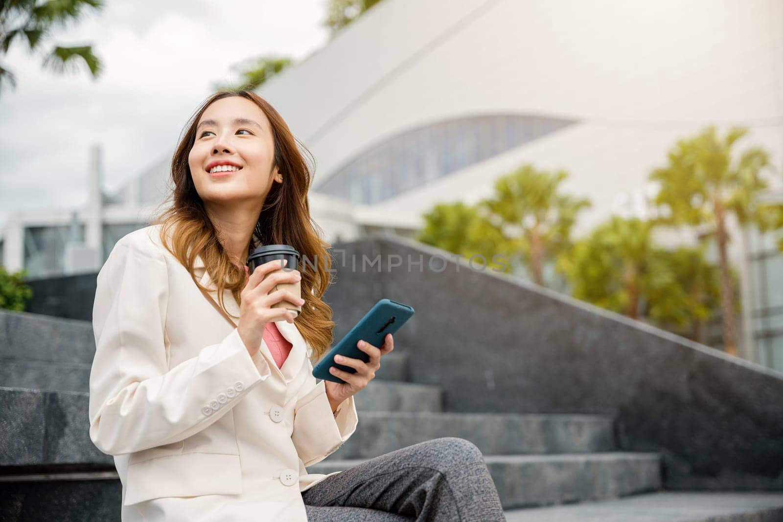 Lifestyle woman business people smiling using mobile smart phones in city office building. Young Asian female wearing blue shirt while use cellphone with holding coffee cup sitting on stair outdoors