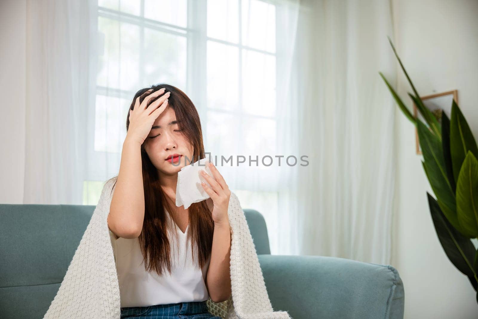 Sick woman sitting under blanket on sofa and sneeze with tissue paper in living room, Asian young female blowing nose sneezing in tissue at home, fever caught cold
