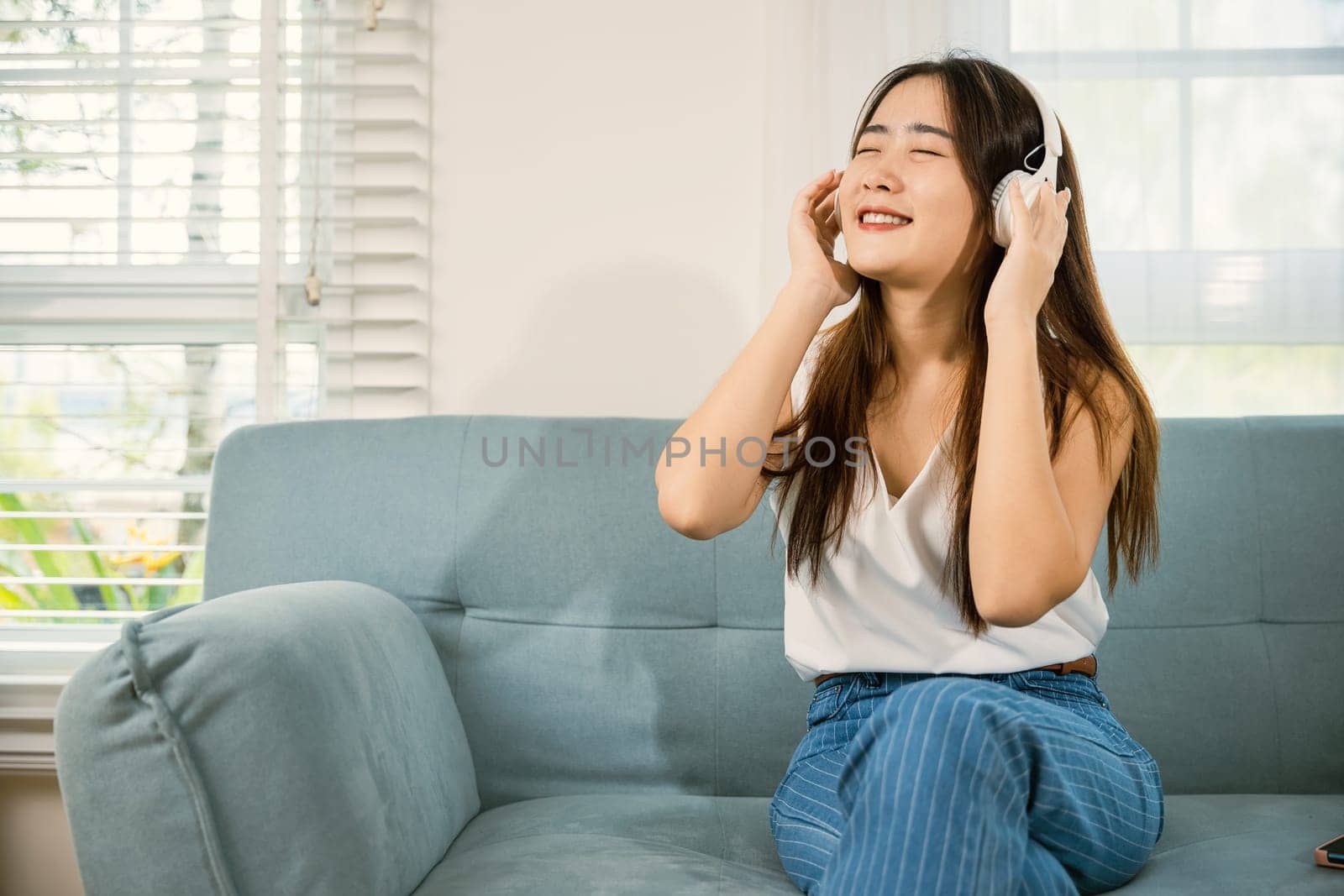 Woman wear earphone listen music application on smartphone, Asian young female wearing headphones listening to music with smart mobile phone and relaxing while sitting on sofa in living room at home