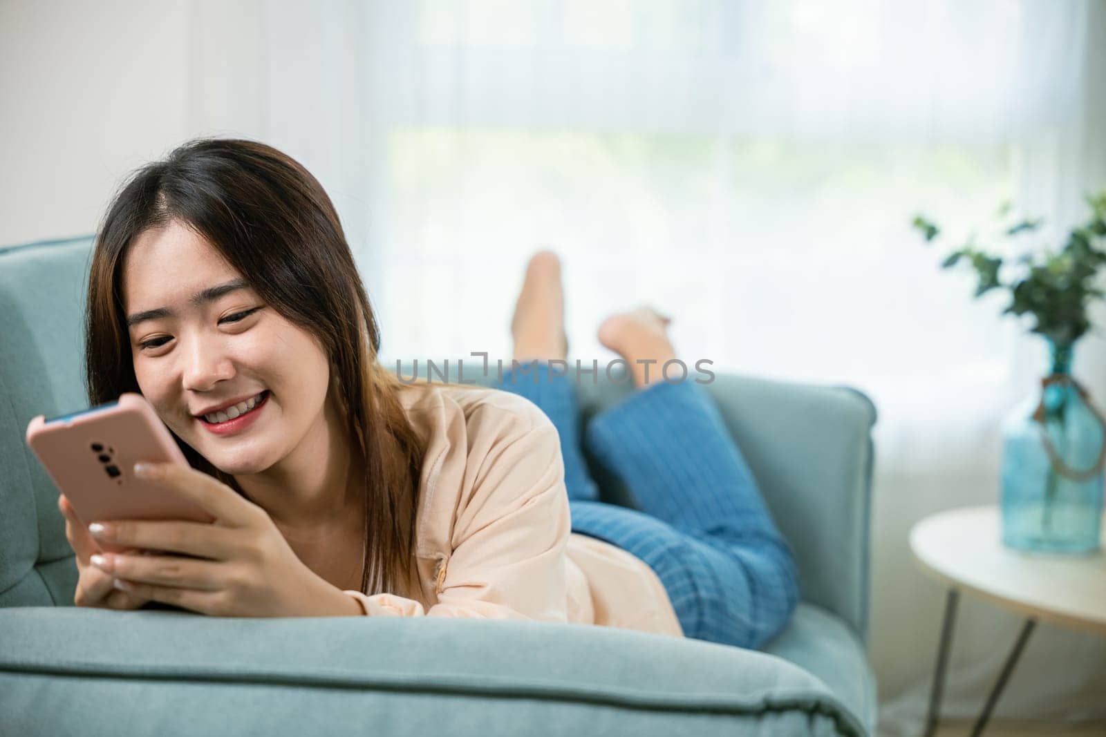 Smiling woman relaxed social online communication on smartphone, Happy Asian female using smart mobile phone lying on sofa in living room room at house, technology gadget online application