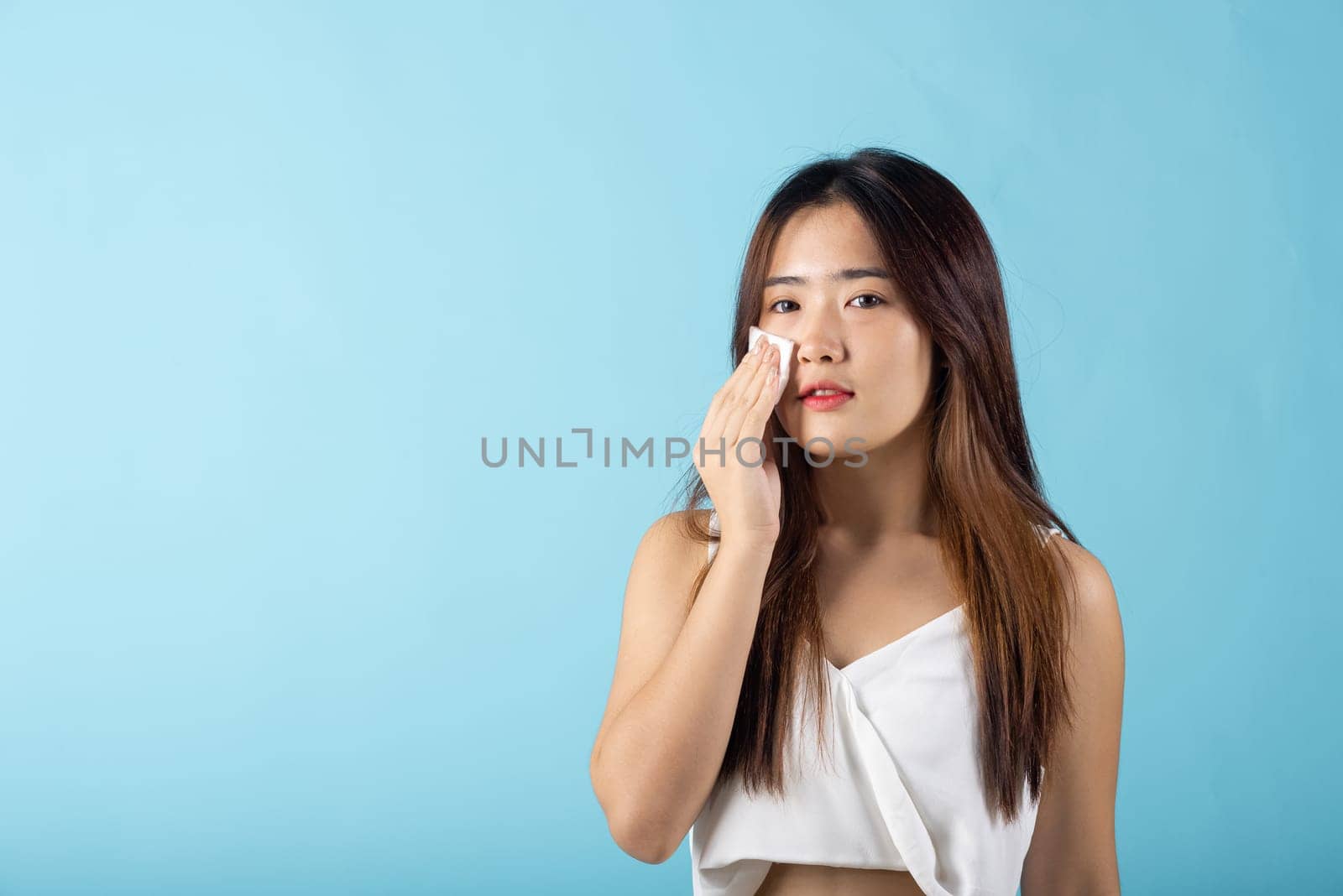 Facial Beauty Treatment. Asian beautiful young woman removing cosmetic make up using pad cotton face cleaner disc, studio shot isolated on blue background, Portrait of happy female cleaning face