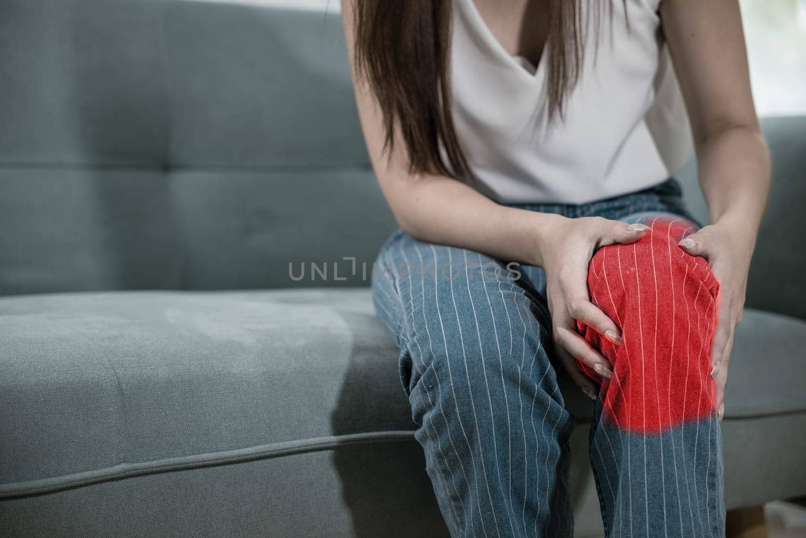Asian young woman sitting on sofa and feeling suffering knee pain in living room at home, female massage her knee leg pain with painful face, Health bone problems self physiotherapy