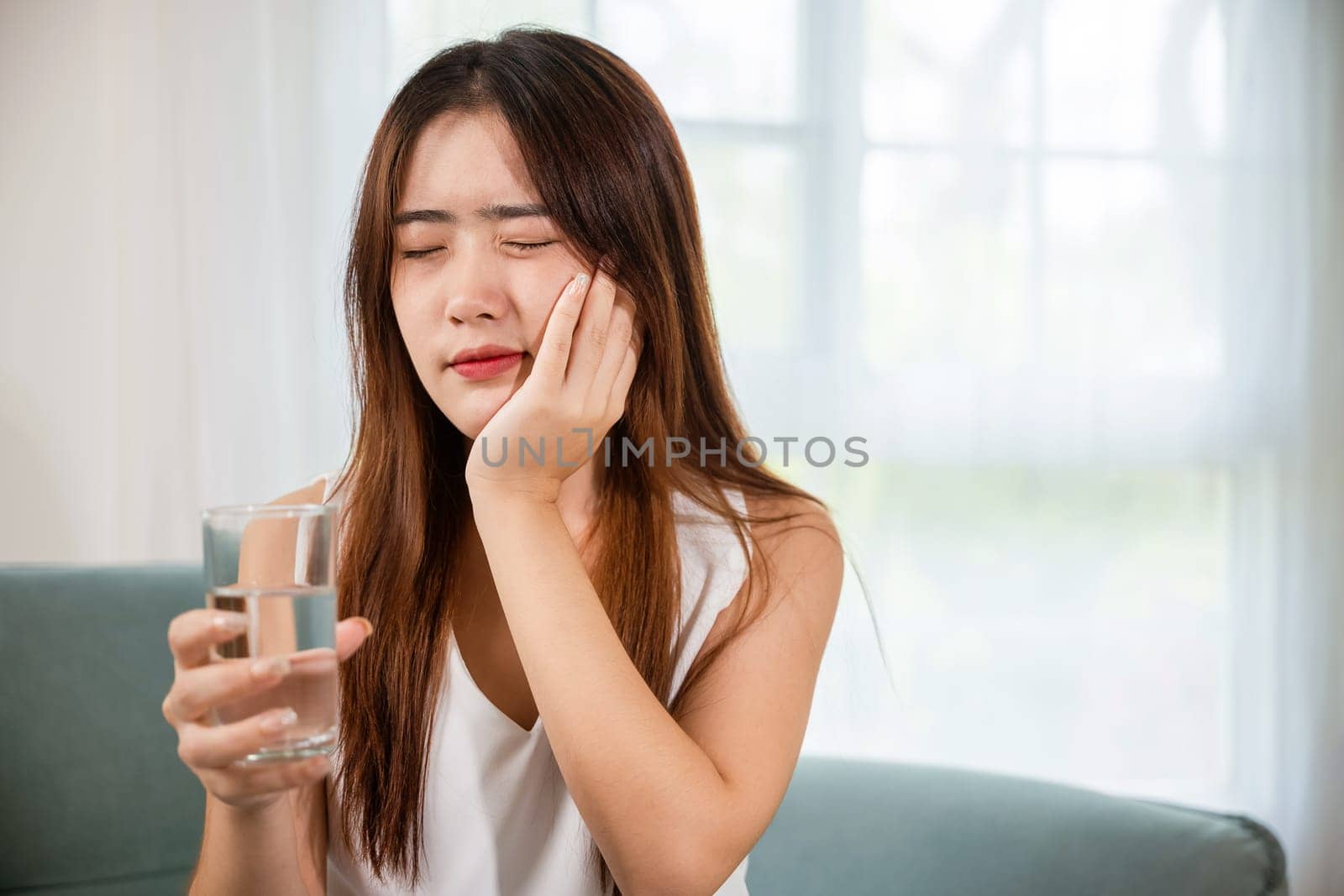 Beautiful female toothache and dental problems touching cheek feeling pain after drinking cold water, Asian young woman with sensitive teeth holding glass of cold water at home in living room, pain
