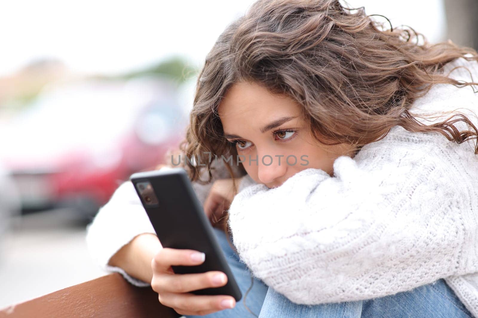 Sad woman checking smart phone sitting in a bench in the street