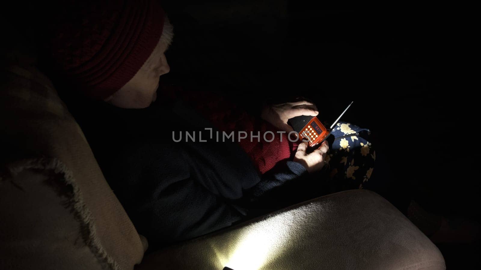 Ukraine: Grandmother holds radio in hands sitting in darkness blackout during russian terroristic attacks. Senior woman in winter clothes in darkness on sofa with hand radio receiver listening news.
