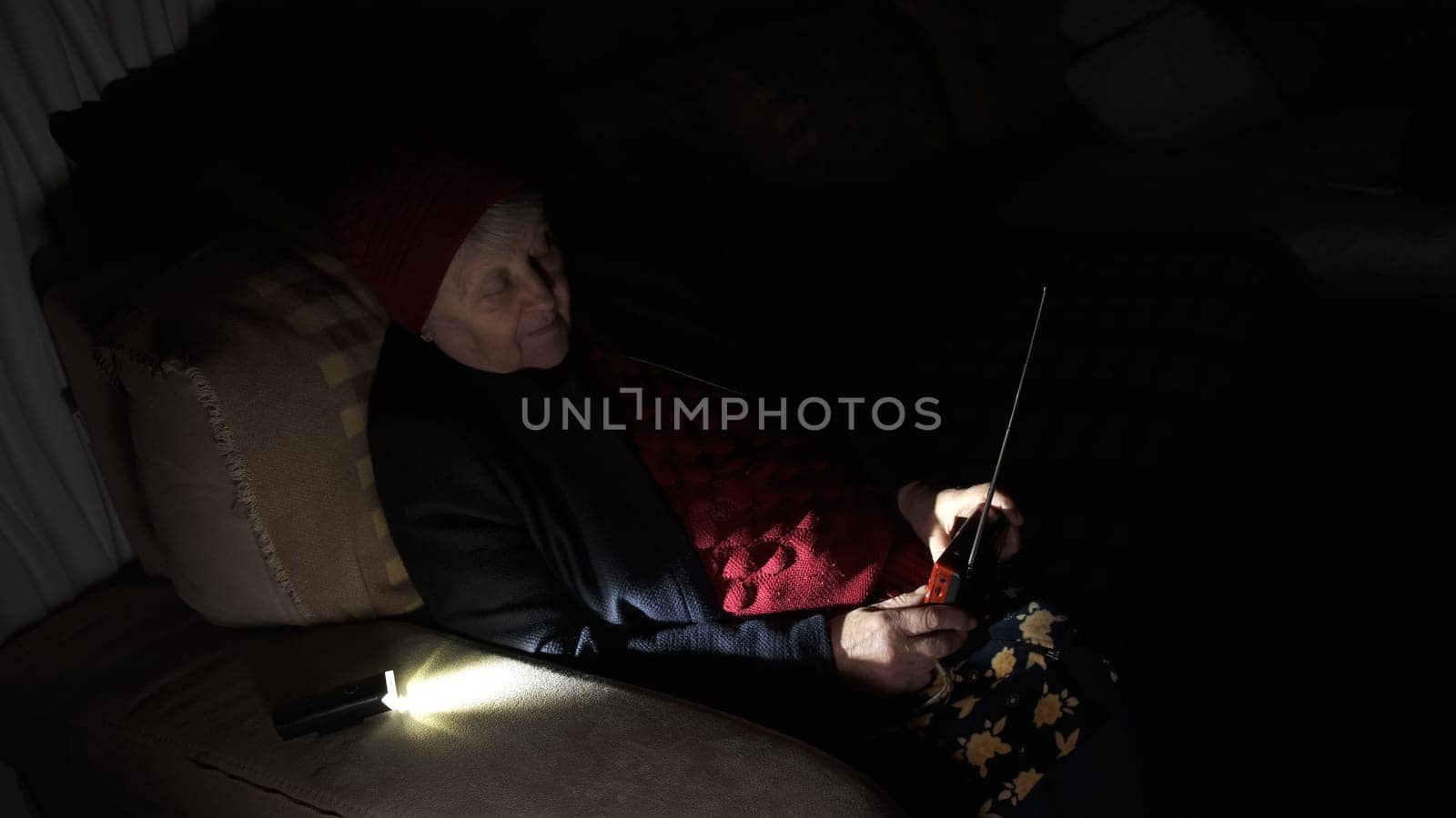 Ukraine: Grandmother holds radio in hands sitting in darkness blackout during russian terroristic attacks. Senior woman in winter clothes in darkness on sofa with hand radio receiver listening news.