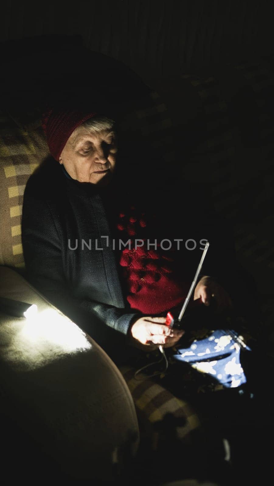 Ukraine: Grandmother holds radio in hands sitting in darkness blackout during russian terroristic attacks. Senior woman in winter clothes in darkness on sofa with hand radio receiver listening news.
