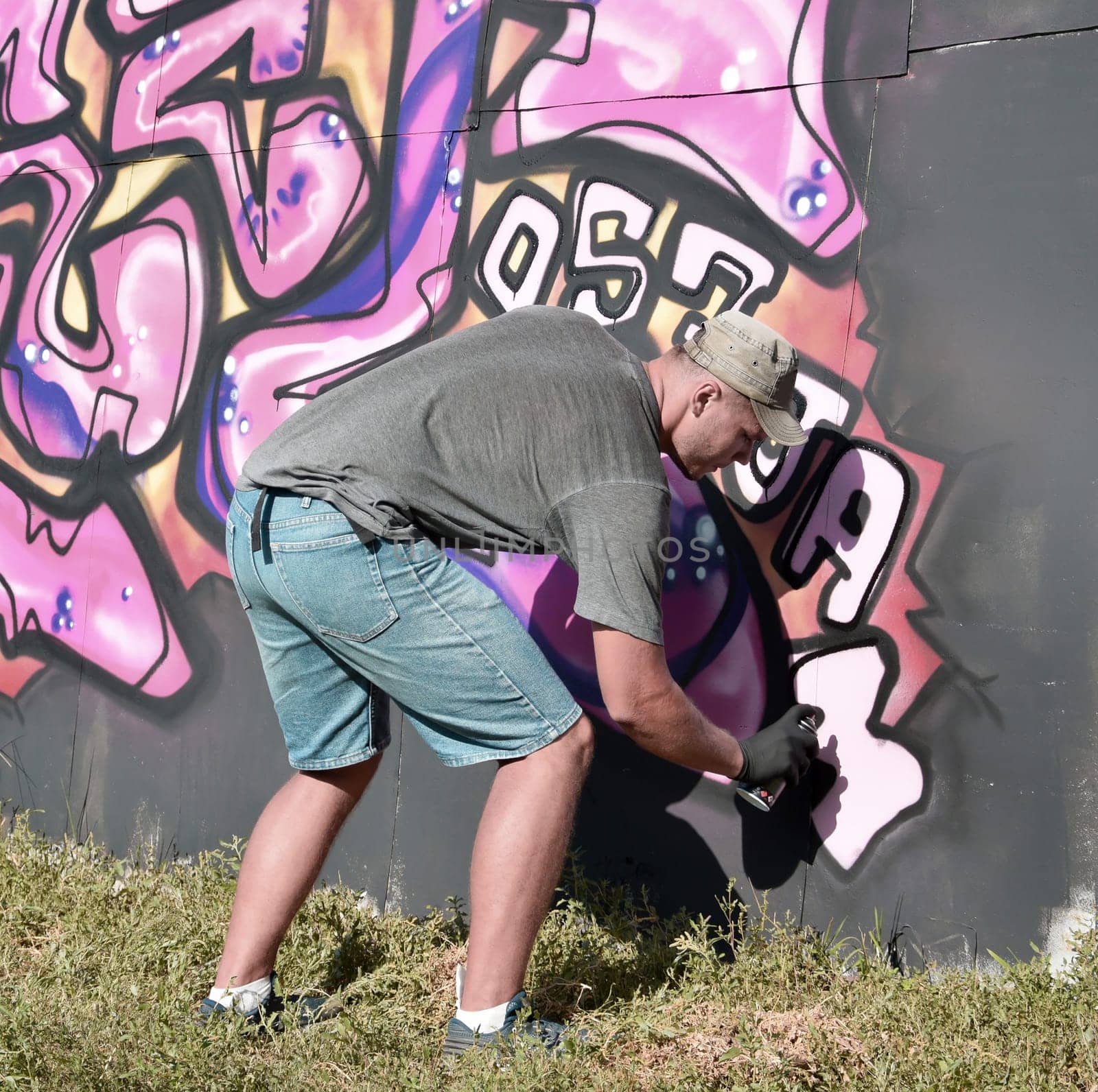Young caucasian male graffiti artist drawing big street art painting in blue and pink tones. Stylish man in denim shorts and grey t shirt made new graffitti artwork outdoors in bright sunny day