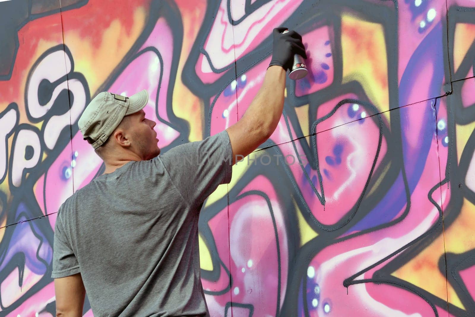 Young caucasian male graffiti artist drawing big street art painting in blue and pink tones. Stylish man in denim shorts and grey t shirt made new graffitti artwork outdoors in bright sunny day