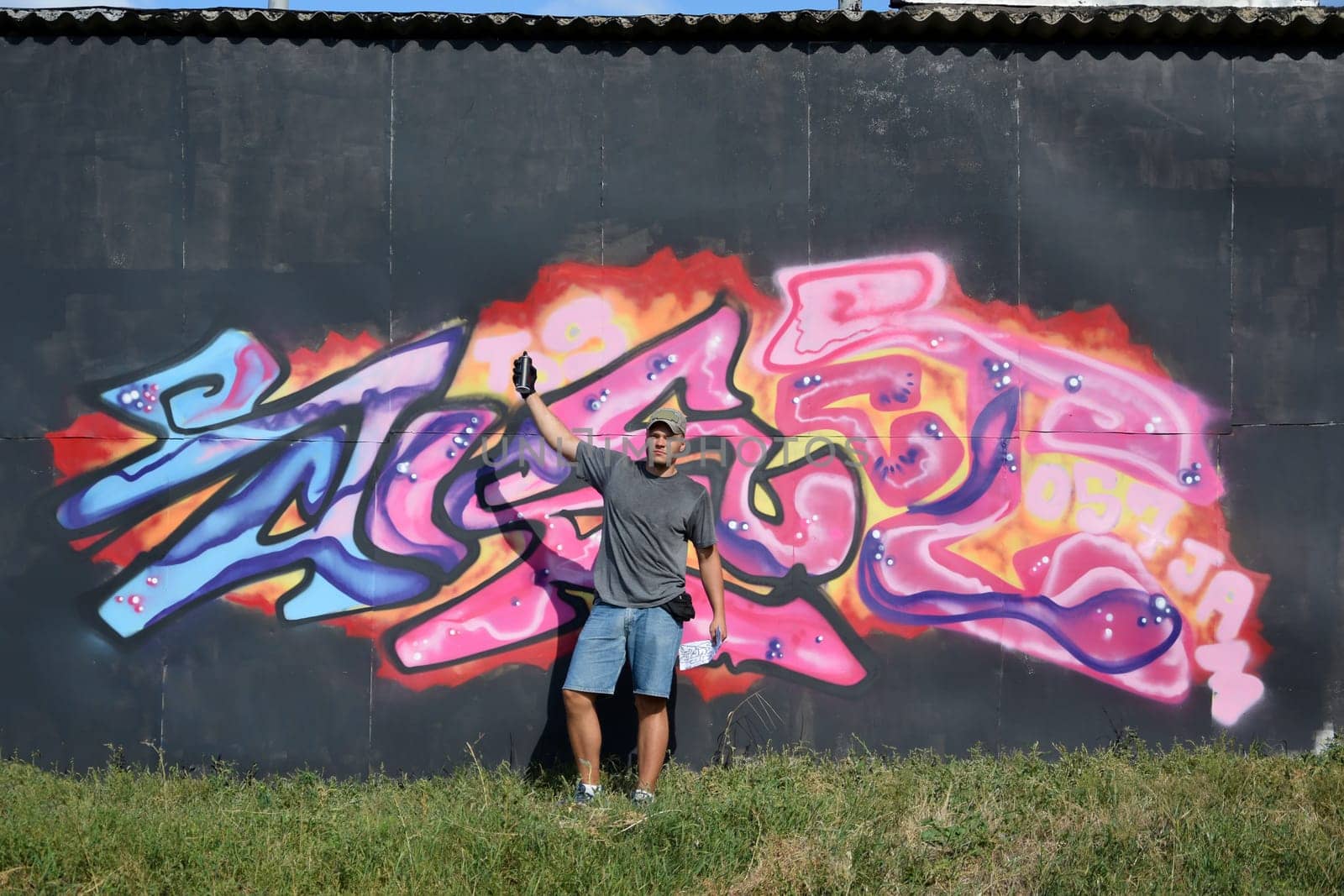 Young caucasian male graffiti artist drawing big street art painting in blue and pink tones. Stylish man in denim shorts and grey t shirt made new graffitti artwork outdoors in bright sunny day