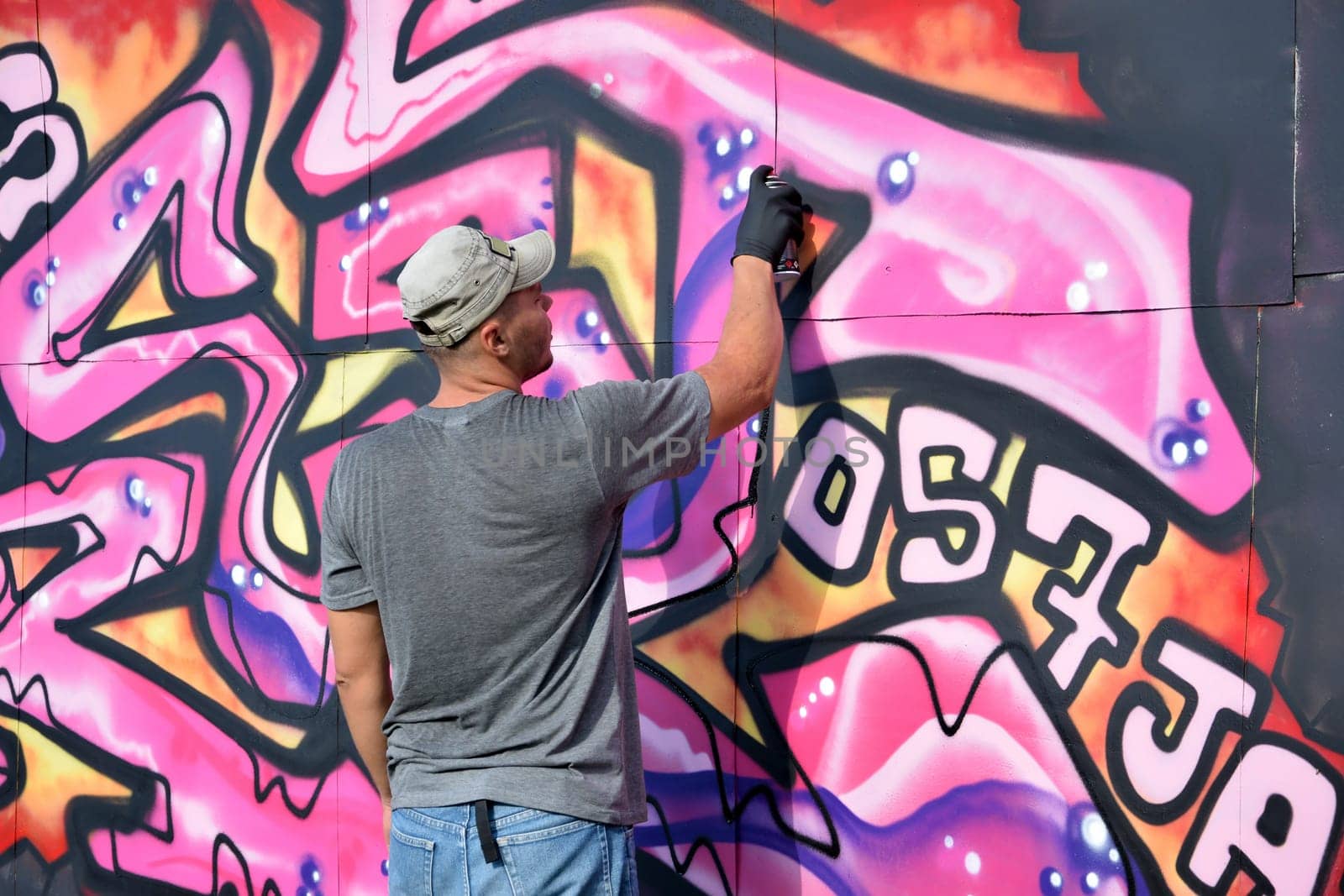 Young caucasian male graffiti artist drawing big street art painting in blue and pink tones. Stylish man in denim shorts and grey t shirt made new graffitti artwork outdoors in bright sunny day