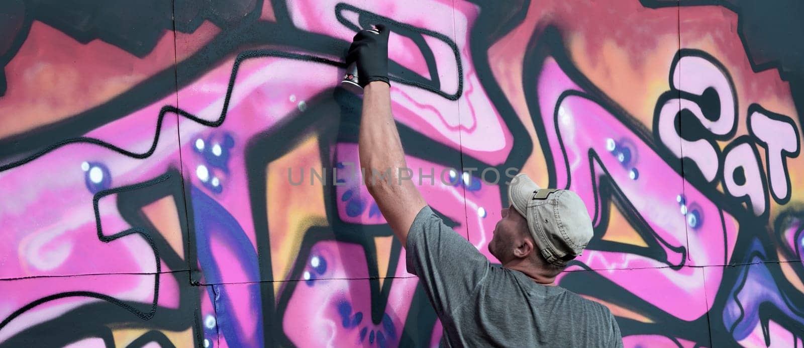 Young caucasian male graffiti artist drawing big street art painting in blue and pink tones. Stylish man in denim shorts and grey t shirt made new graffitti artwork outdoors in bright sunny day