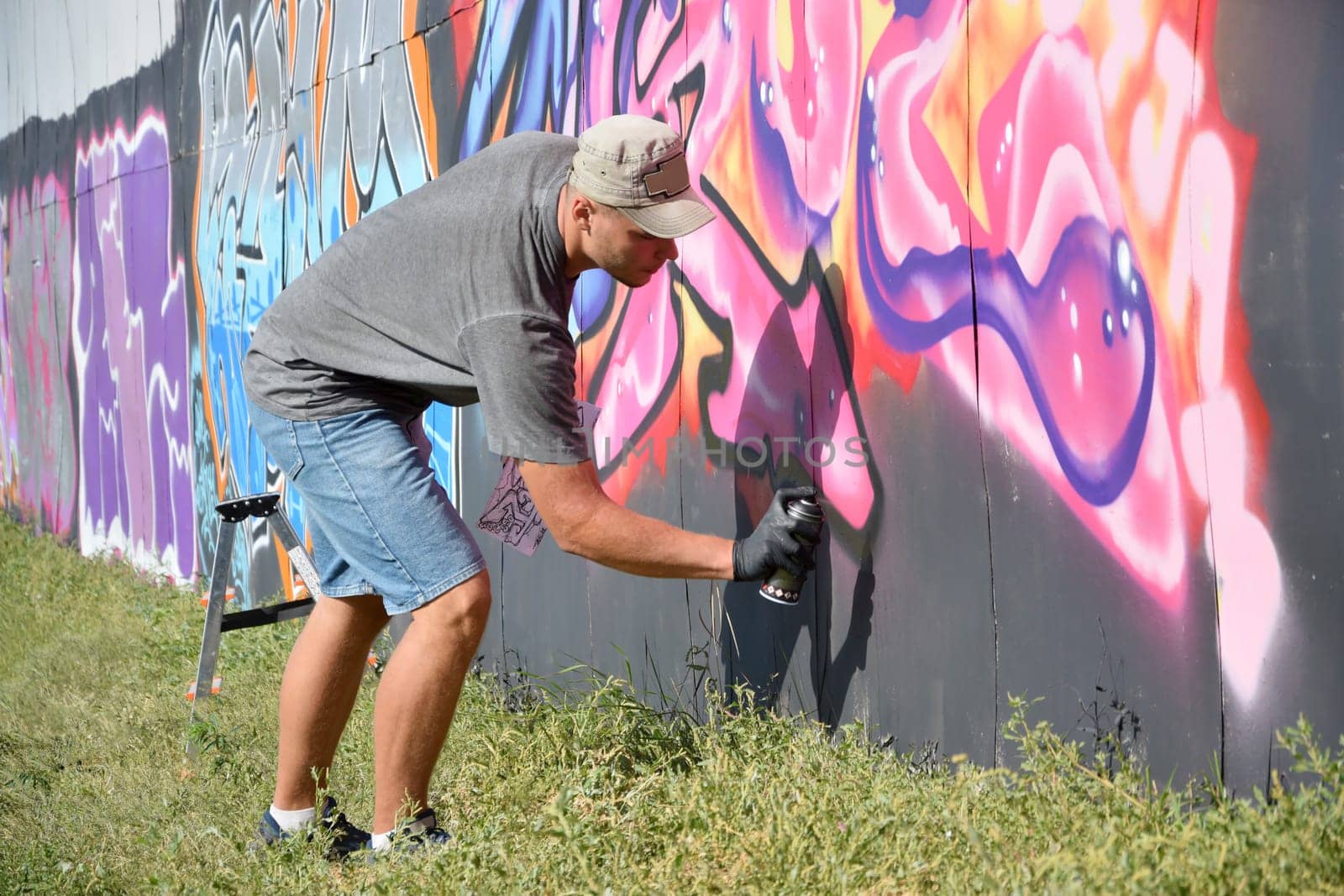Young caucasian male graffiti artist drawing big street art painting in blue and pink tones. Stylish man in denim shorts and grey t shirt made new graffitti artwork outdoors in bright sunny day