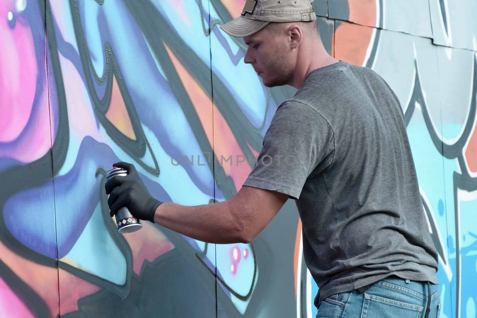 Young caucasian male graffiti artist drawing big street art painting in blue and pink tones. Stylish man in denim shorts and grey t shirt made new graffitti artwork outdoors in bright sunny day