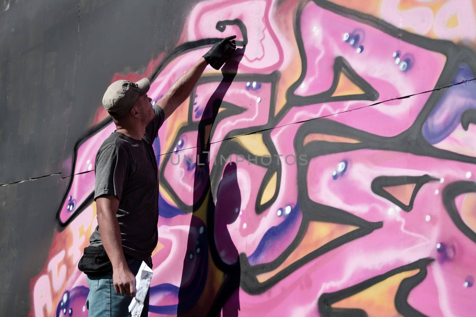 Young caucasian male graffiti artist drawing big street art painting in blue and pink tones. Stylish man in denim shorts and grey t shirt made new graffitti artwork outdoors in bright sunny day
