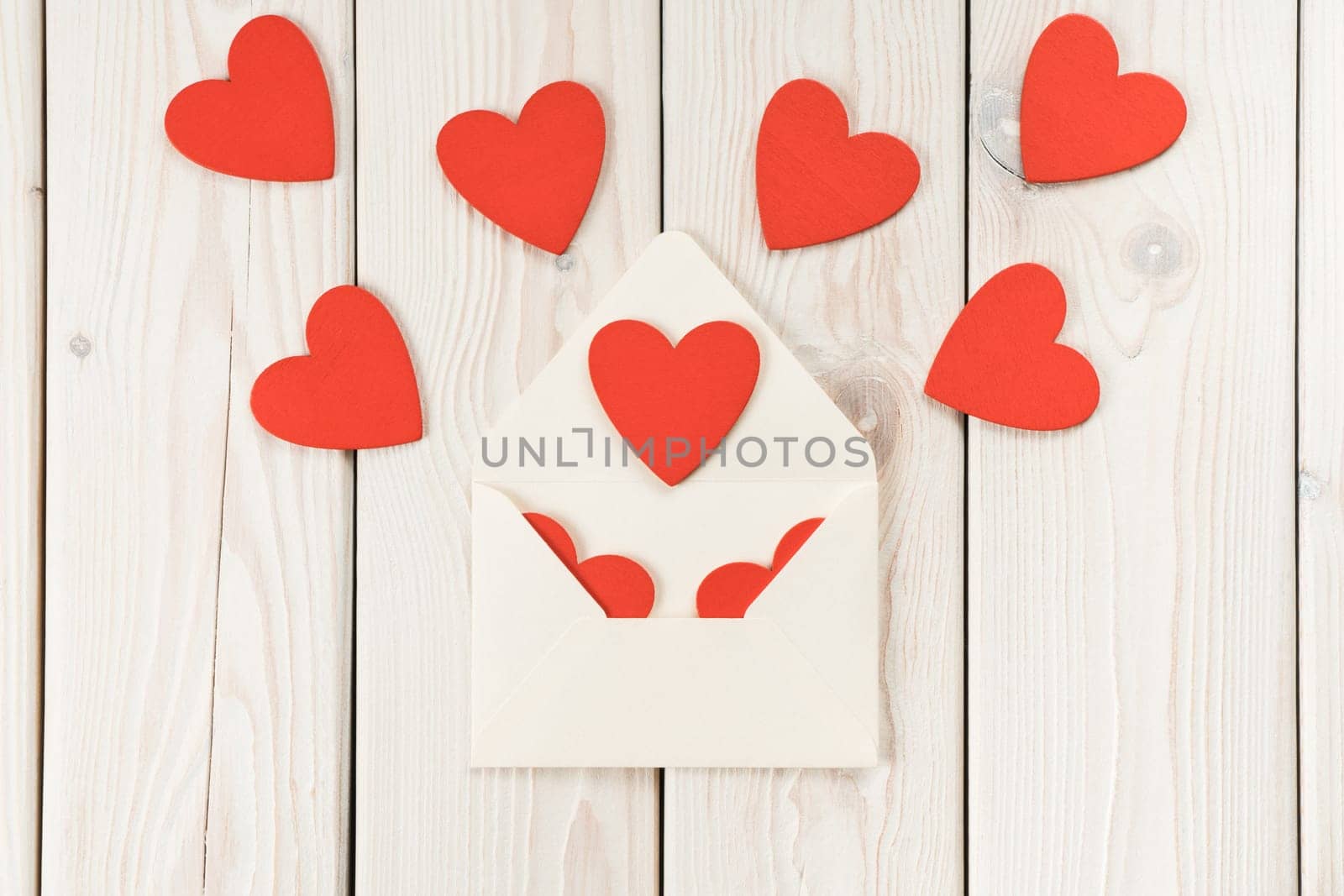 White paper craft envelope opening and giving composition of red paper hearts on light wooden background. St Valentines, women, birthday or wedding celebration message, letter. Copy space, flat lay
