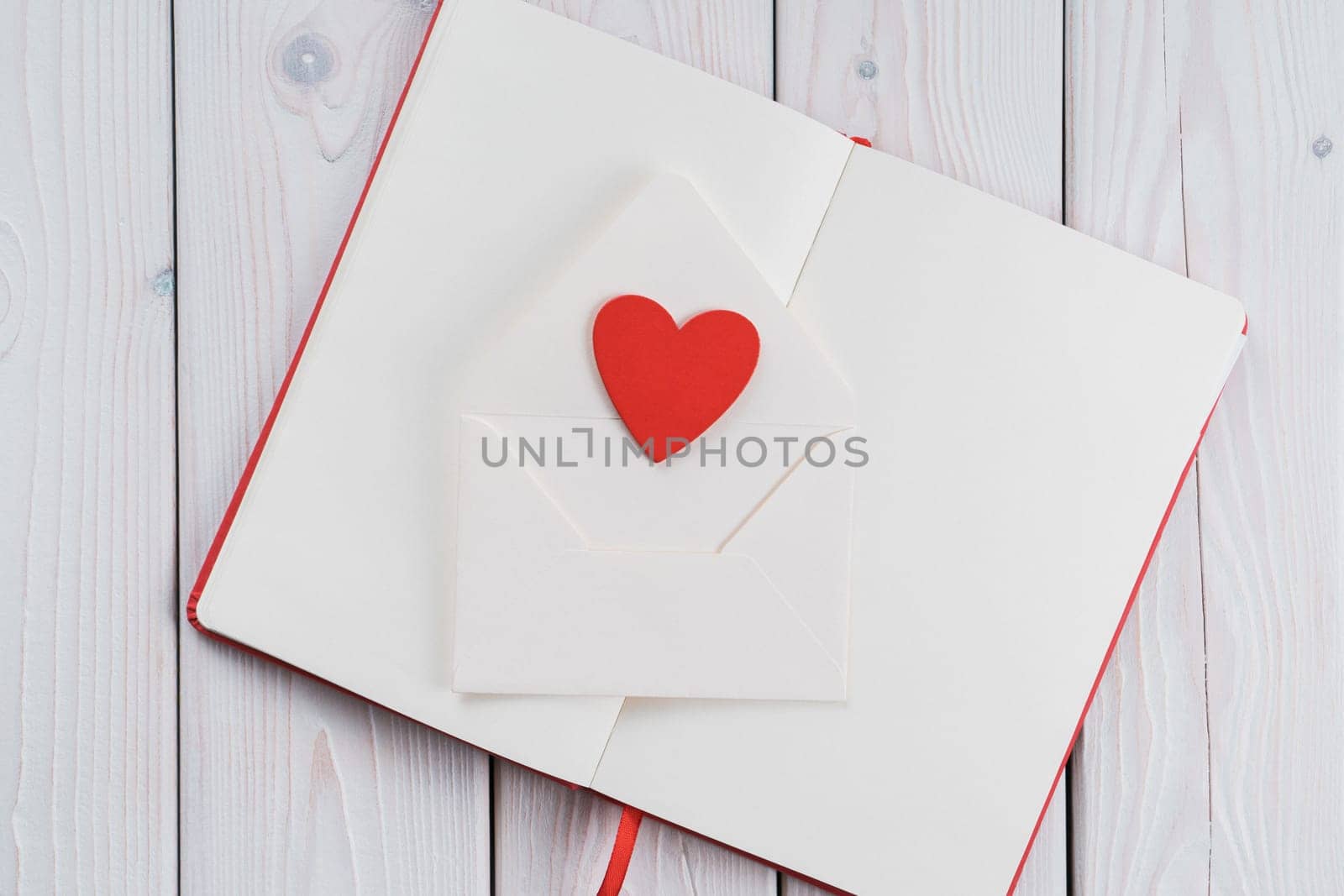 Red diary book with white paper with red heart in craft envelope on light wooden desk background. Sending love letter, message. Falling in love in saint valentines day or 8 march. Copy space, flat lay