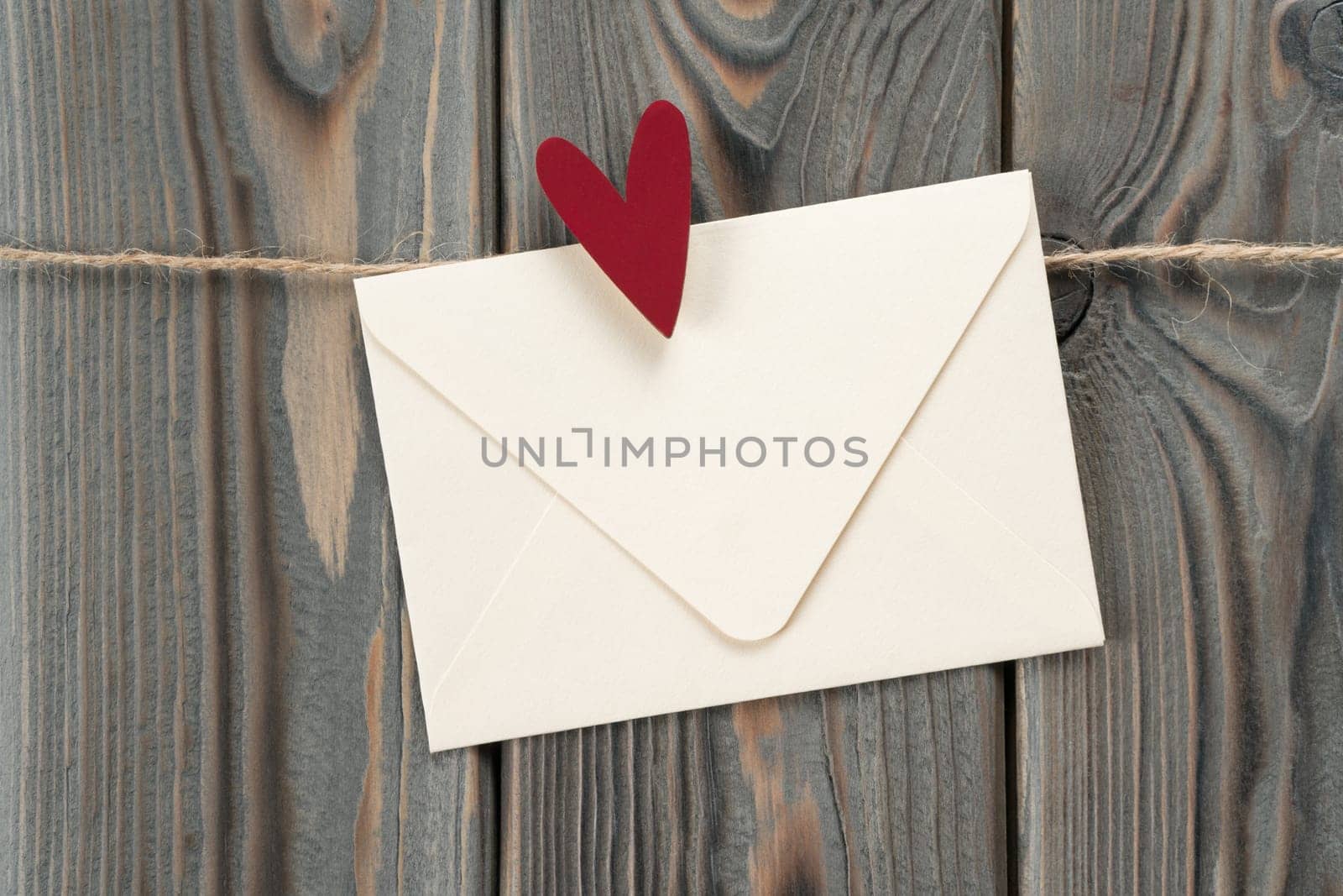 Cute handmade white paper envelope with red heart as stamp, hanging on rope on dark wooden background. Secret present and send feelings on saint Valentine Day. Love letter. Copy space, mockup